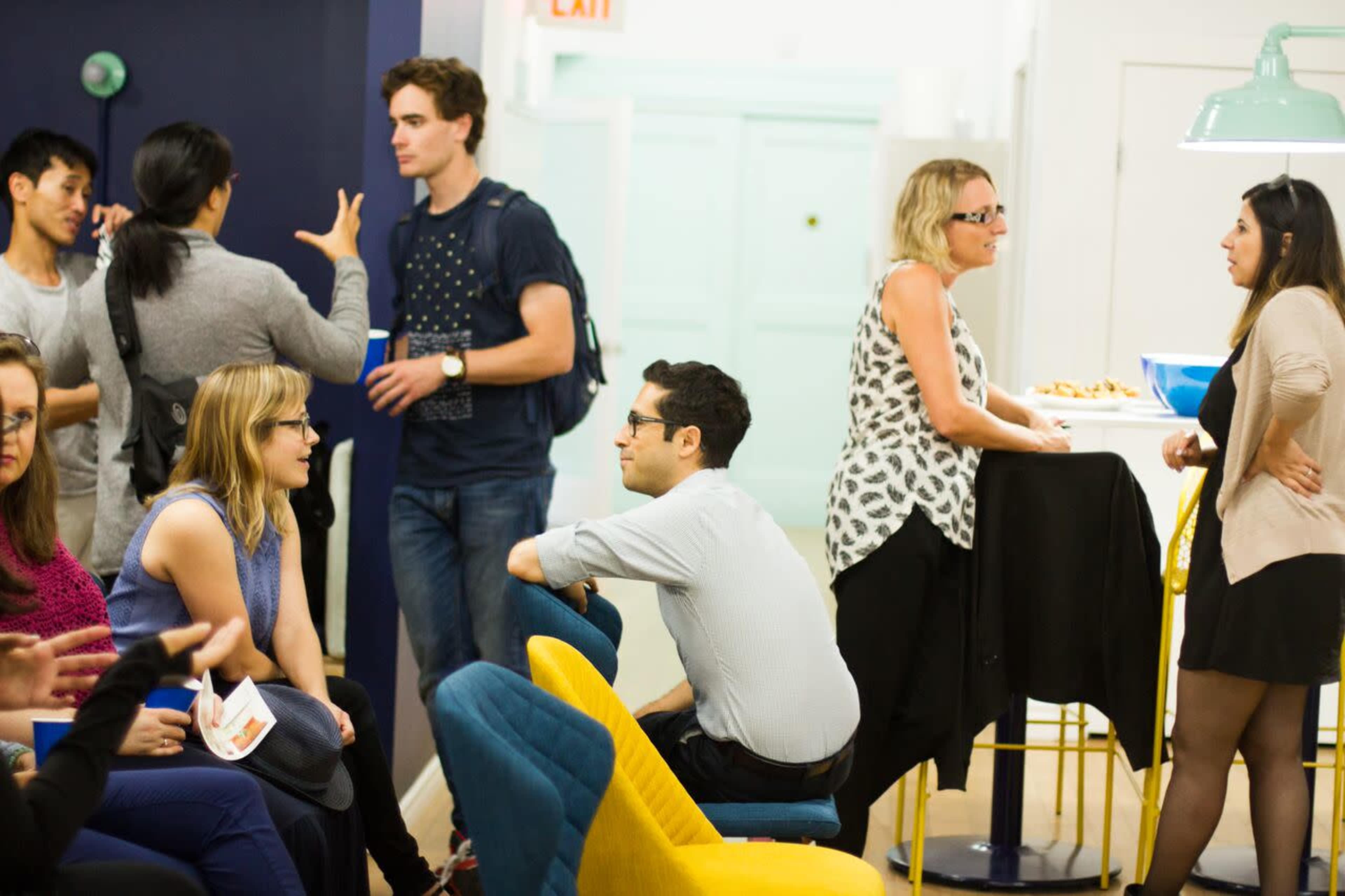 A group of people gathers in a brightly lit room with colorful seating, some engaged in conversation and others seated together.
