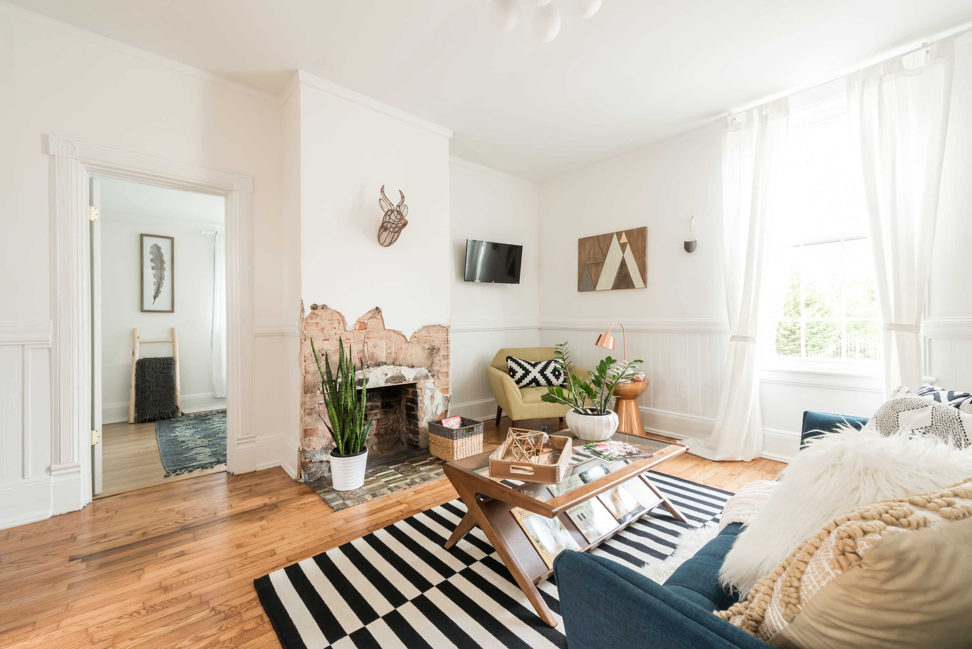 A living room features a mix of furniture with a coffee table, a television mounted on the wall, and large windows allowing natural light to illuminate the space.