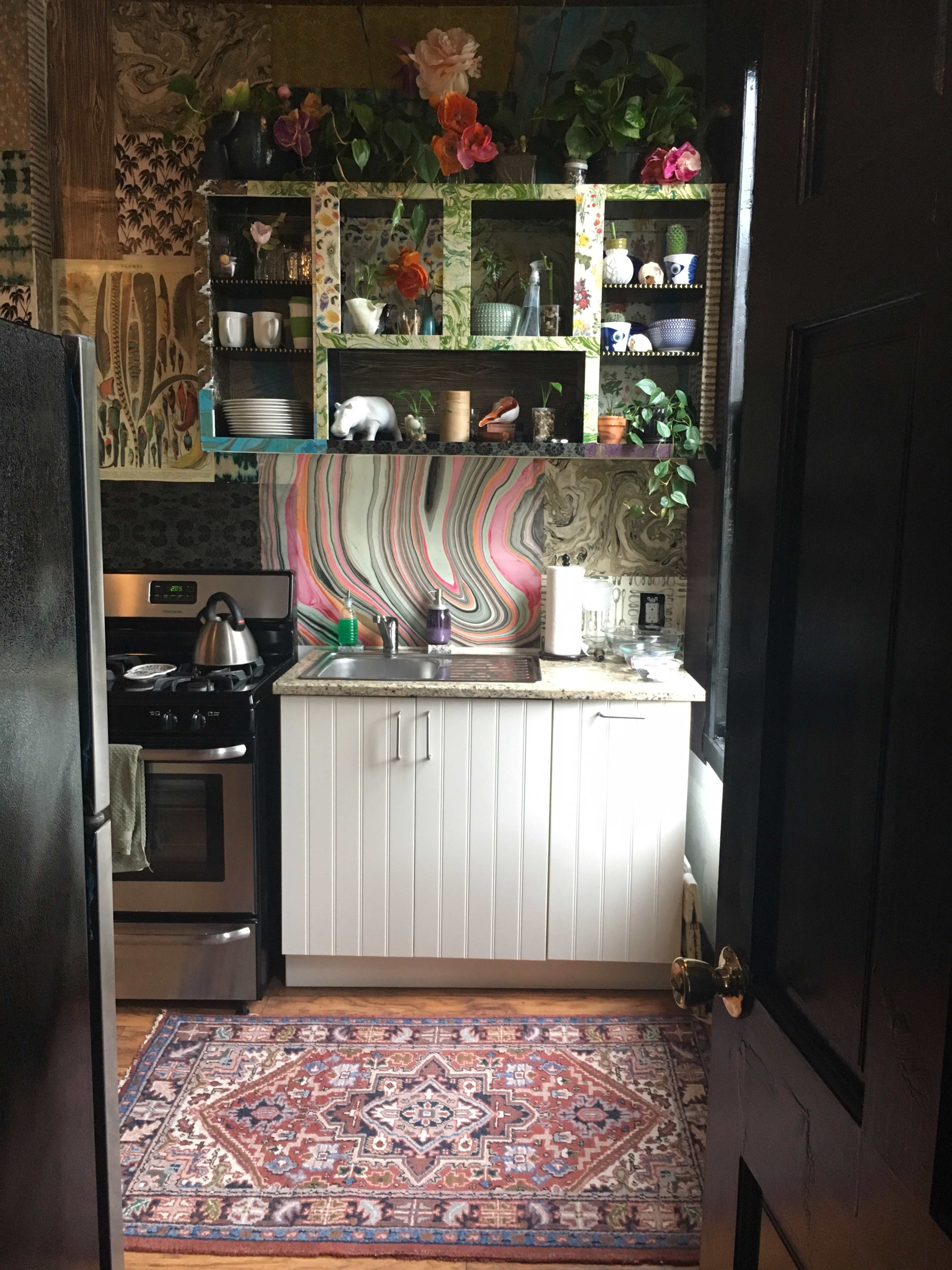A kitchen is visible, featuring a sink, stove, and a patterned rug, with colorful shelves adorned with plants and various objects against a marbled wall.