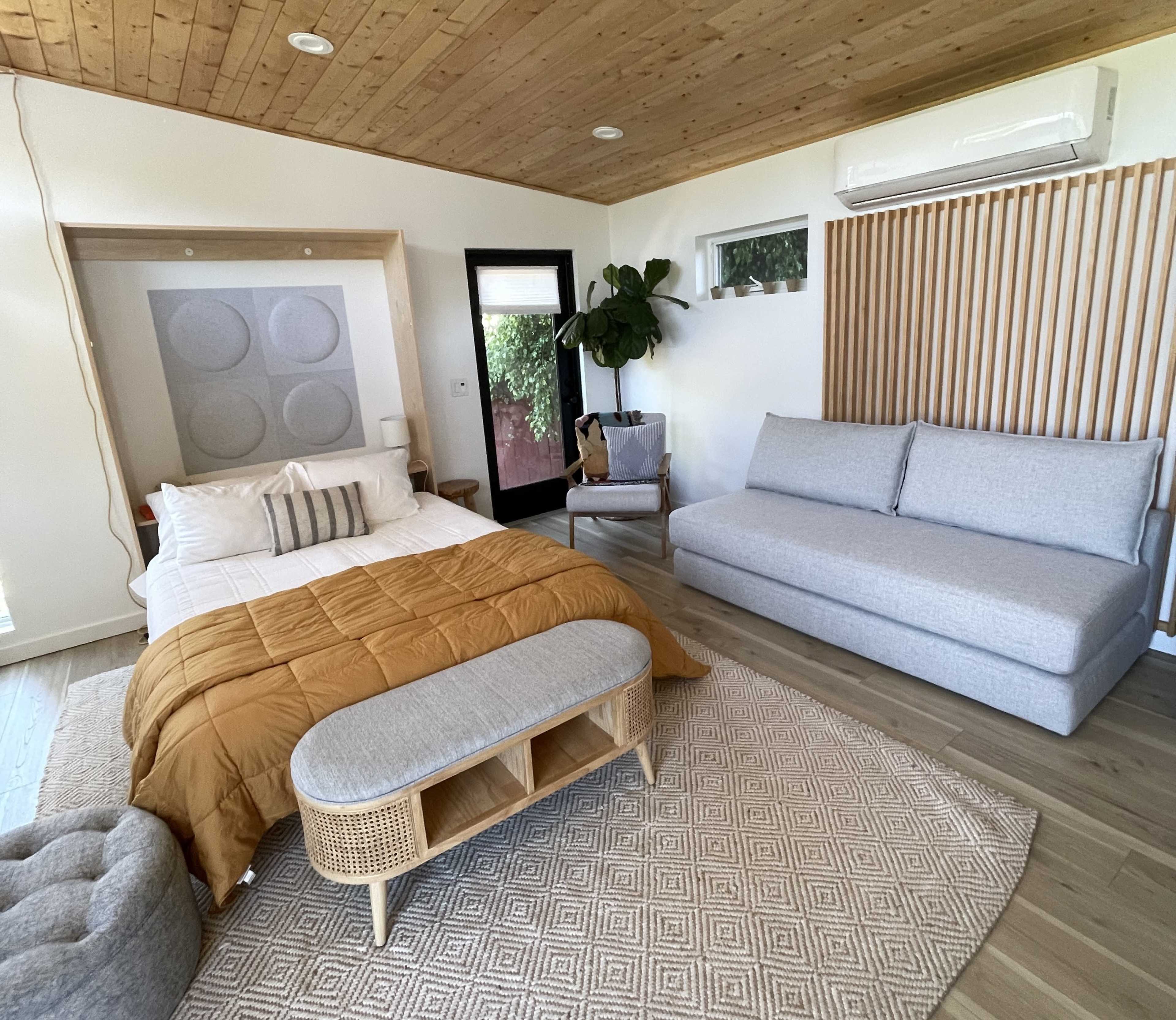 A cozy bedroom featuring a bed with a mustard-colored duvet, a gray sofa, an area rug, and a potted plant, all set against a wooden ceiling and light walls.