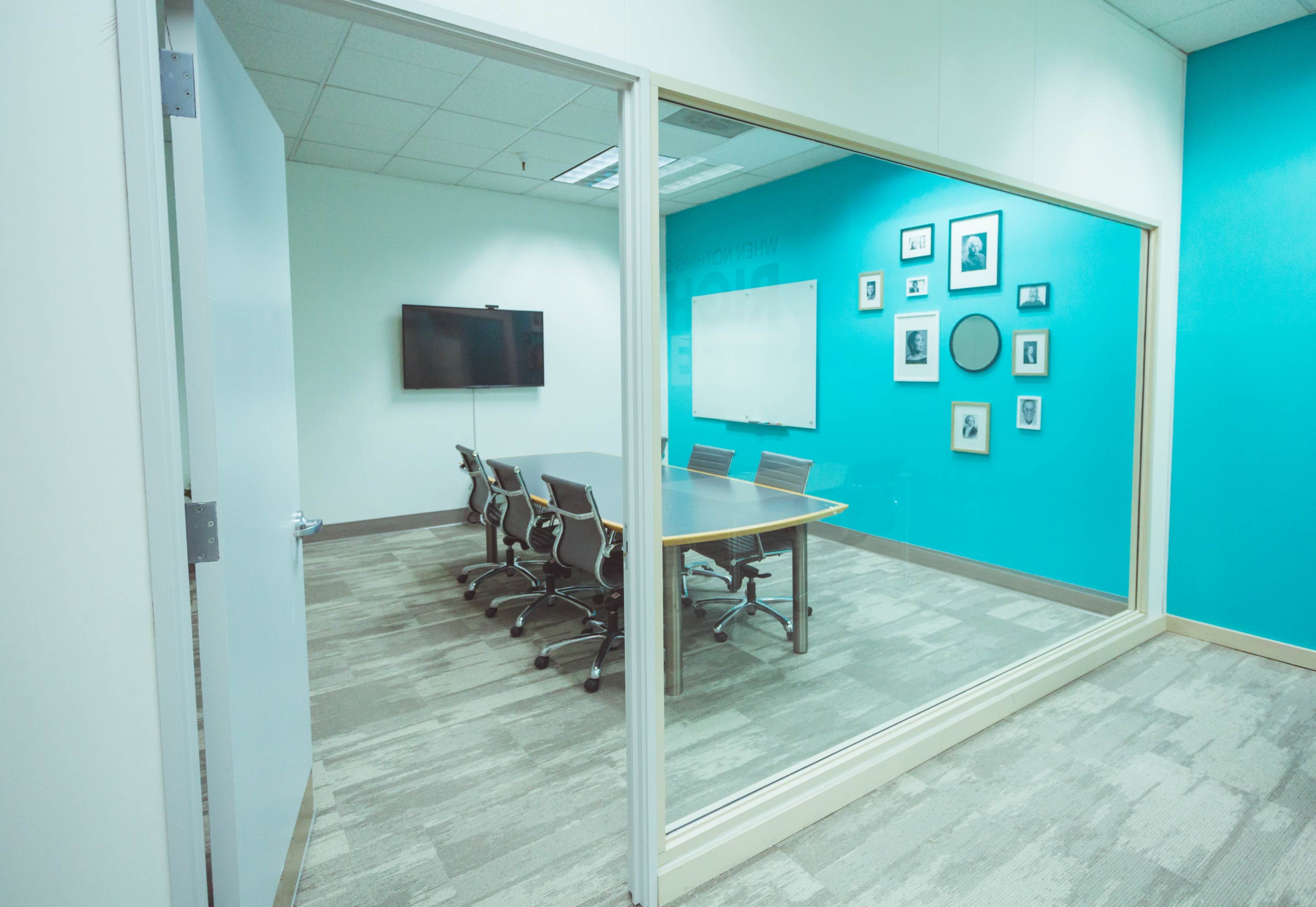 A conference room with a large table and ergonomic chairs, visible through a glass wall, featuring a television and framed photos on the turquoise wall.
