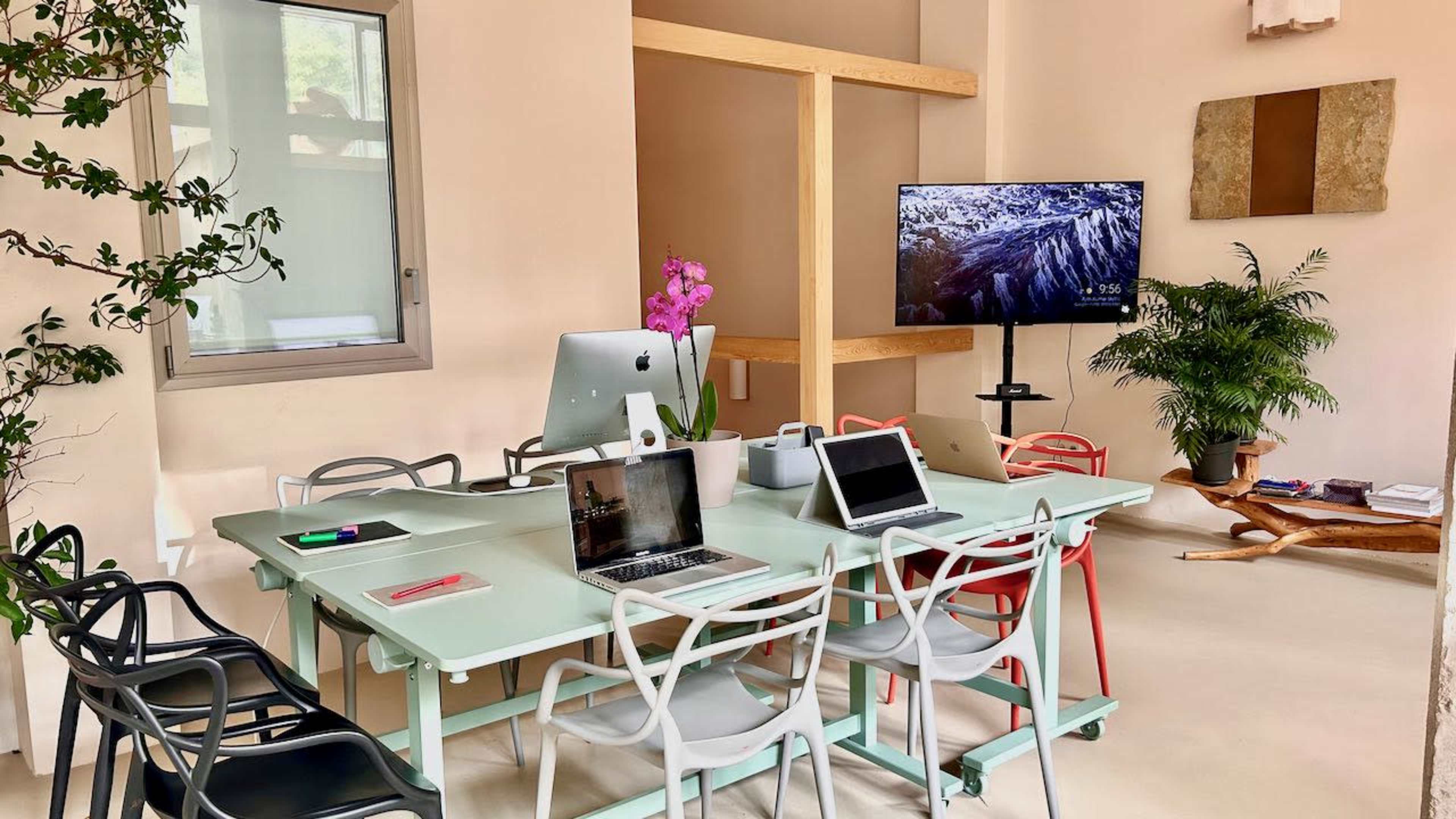 A modern meeting room features a large glass table surrounded by colorful chairs, a TV mounted on the wall, and multiple devices on the table.