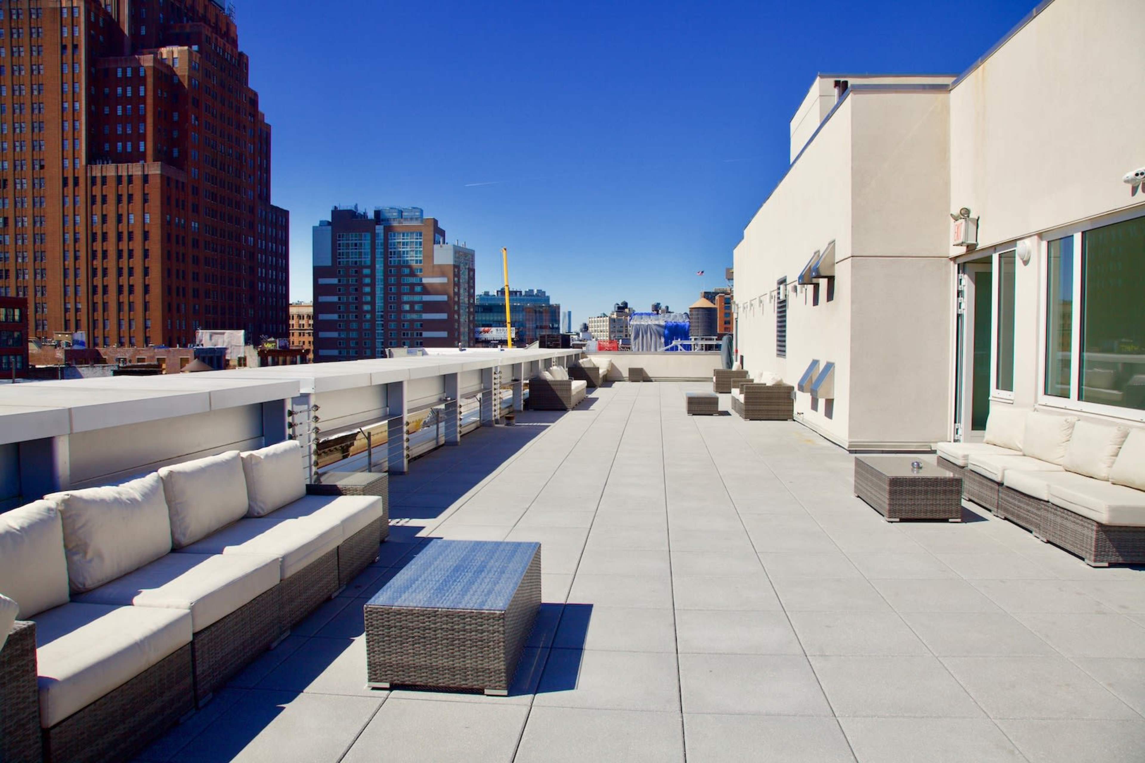 A flat rooftop terrace features modern furniture, including couches and tables, against a backdrop of city buildings under a clear blue sky.