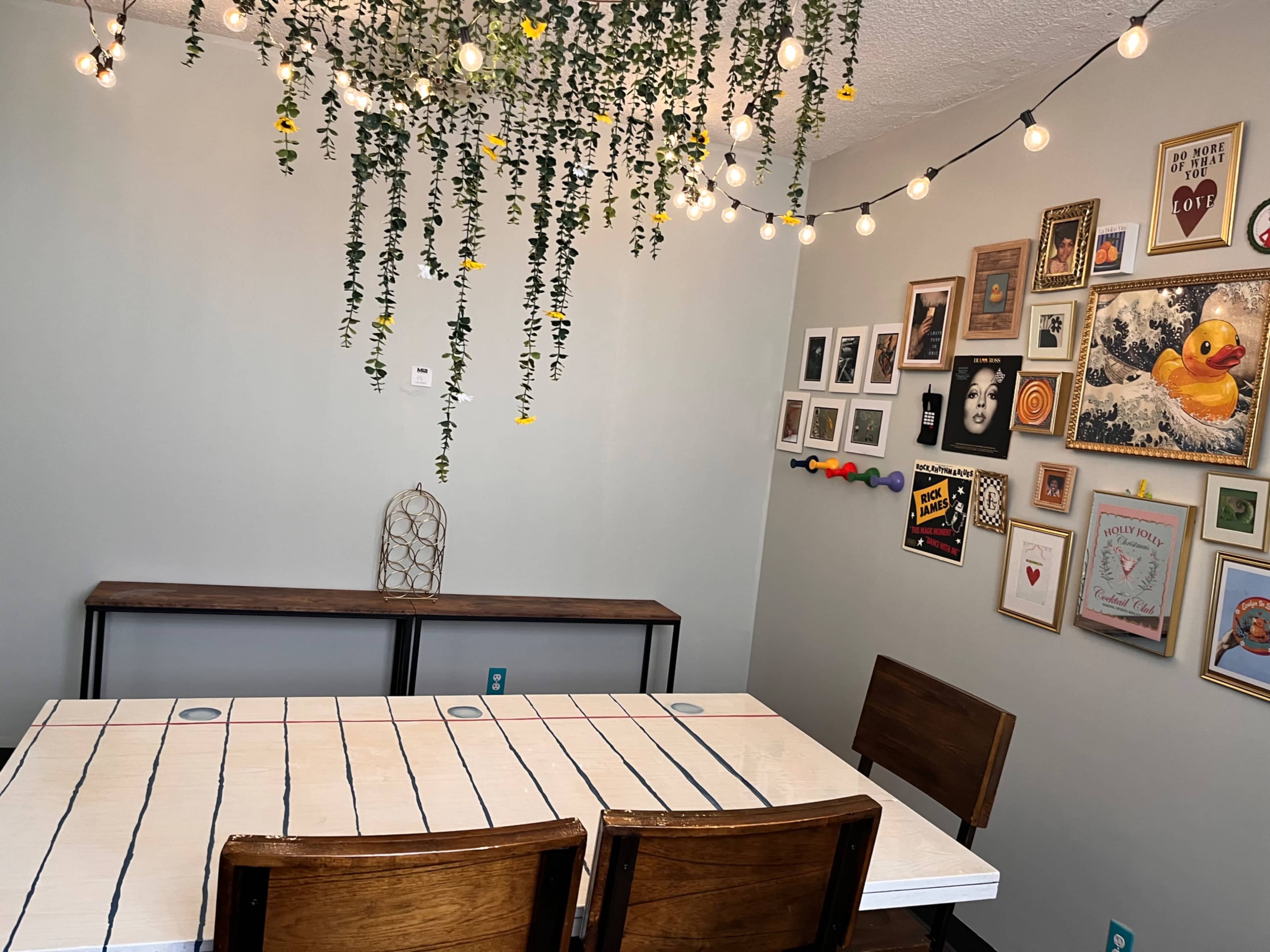 The image shows a dining area with a wooden table and chairs, a wall adorned with various framed artworks, and string lights hanging from the ceiling with greenery.