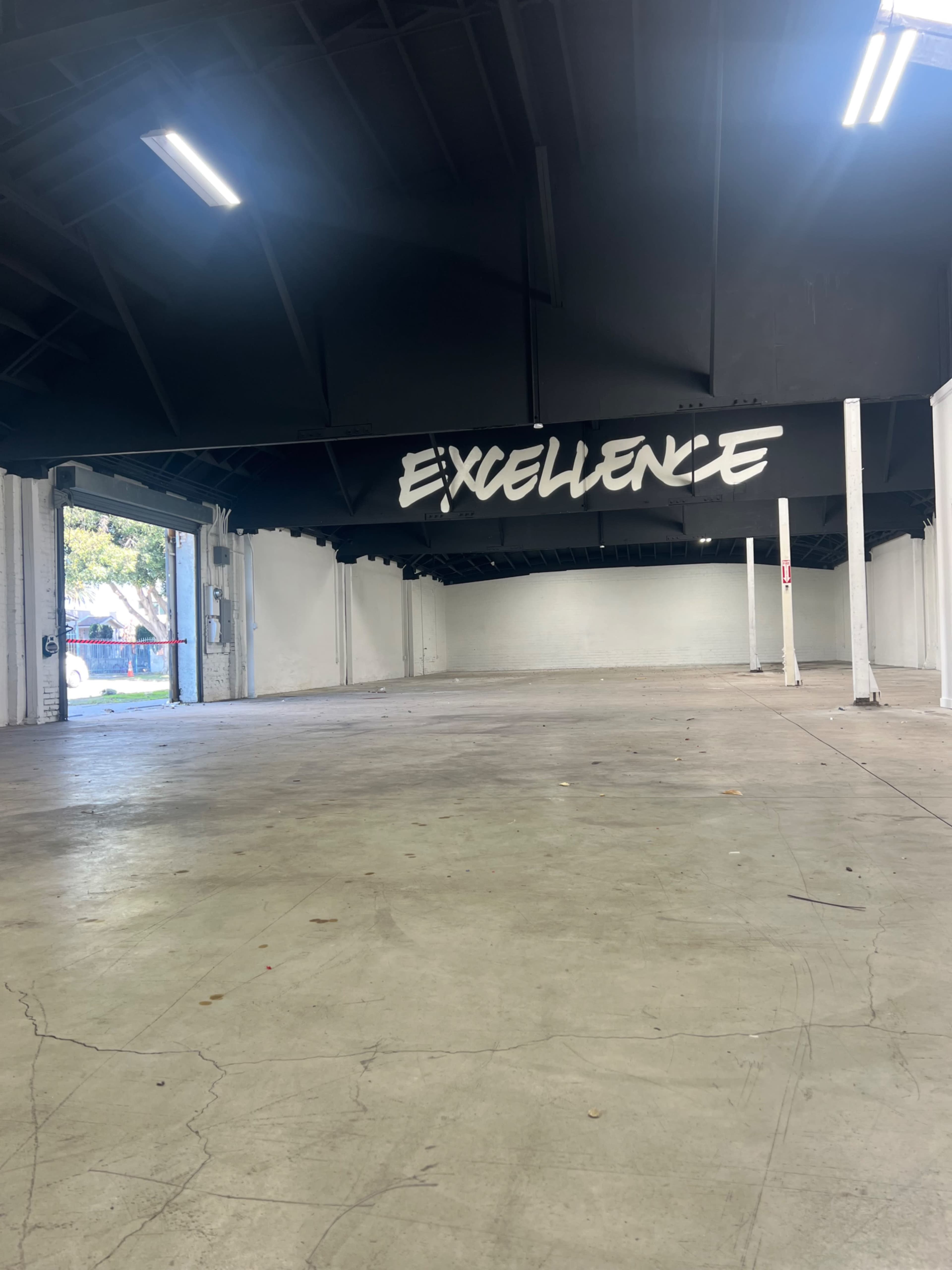 An empty warehouse interior features a large black sign on the ceiling that reads "EXCELLENCE."