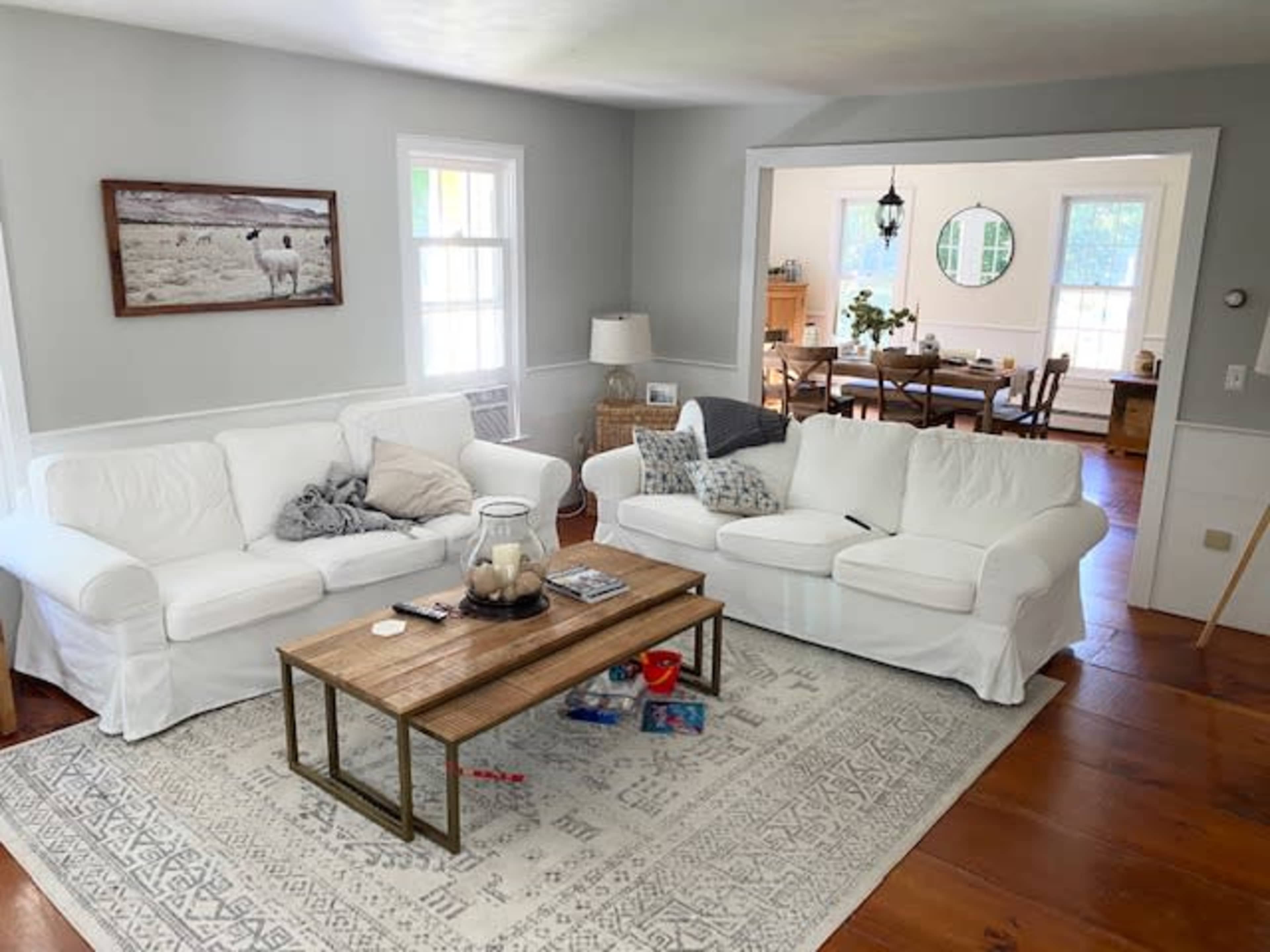 The image shows a living room with white sofas, a wooden coffee table, and a light-colored area rug, leading into a dining area in the background.