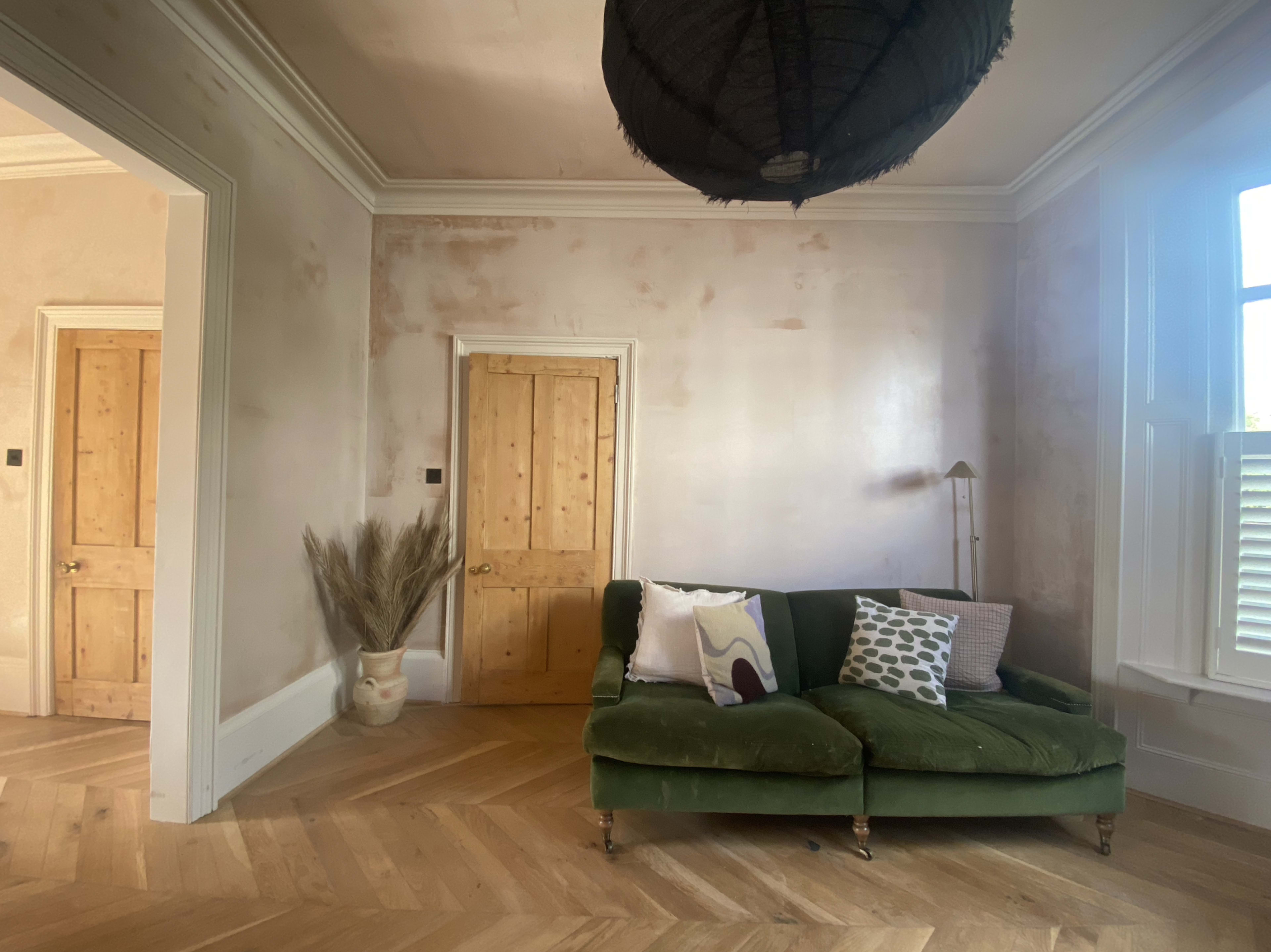 A room with a green velvet sofa facing a pair of wooden doors, with a large woven pendant lamp overhead and a decorative plant in a pot nearby.