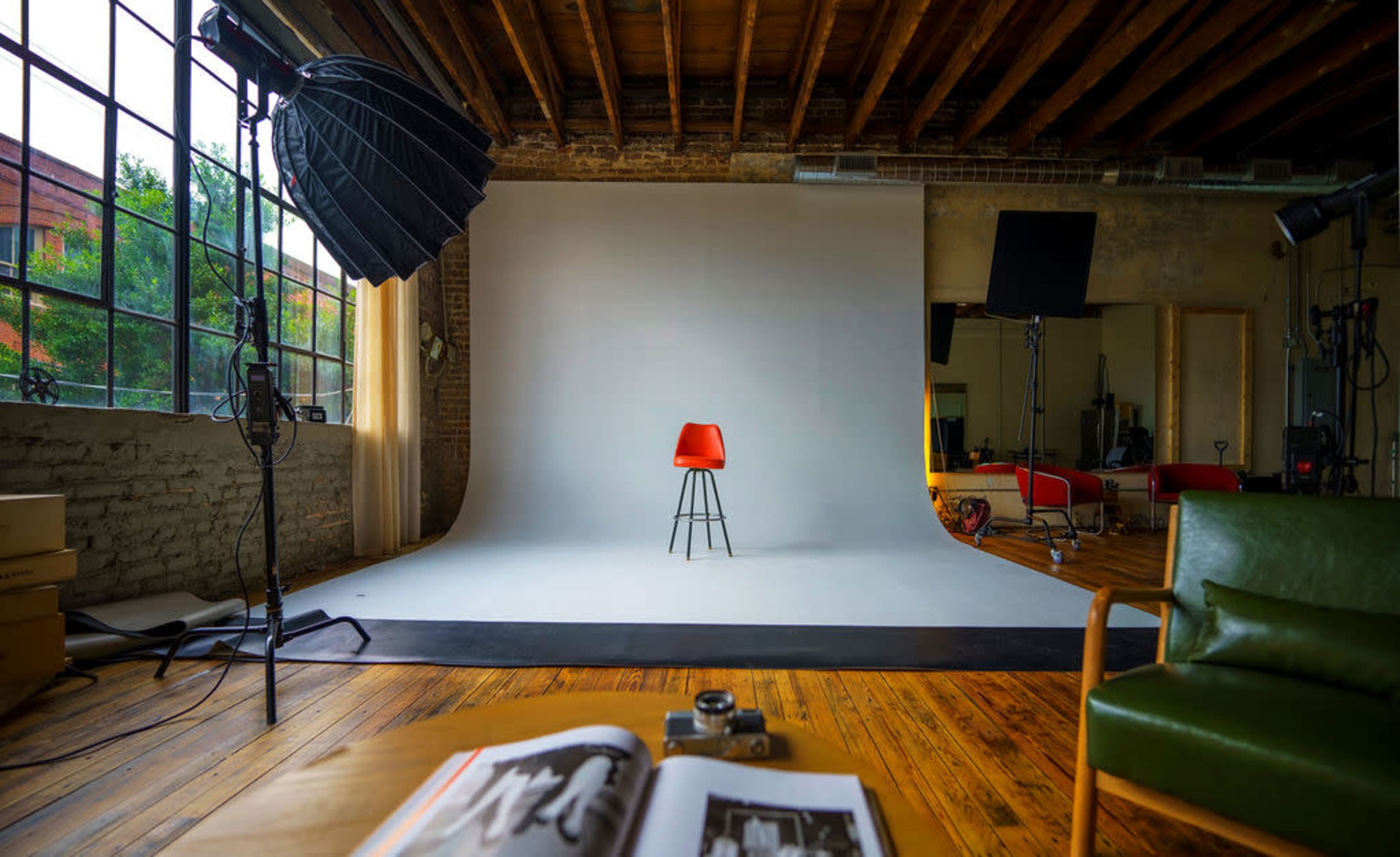The image shows a photography studio featuring a red chair positioned on a white backdrop, surrounded by lights and a large window.