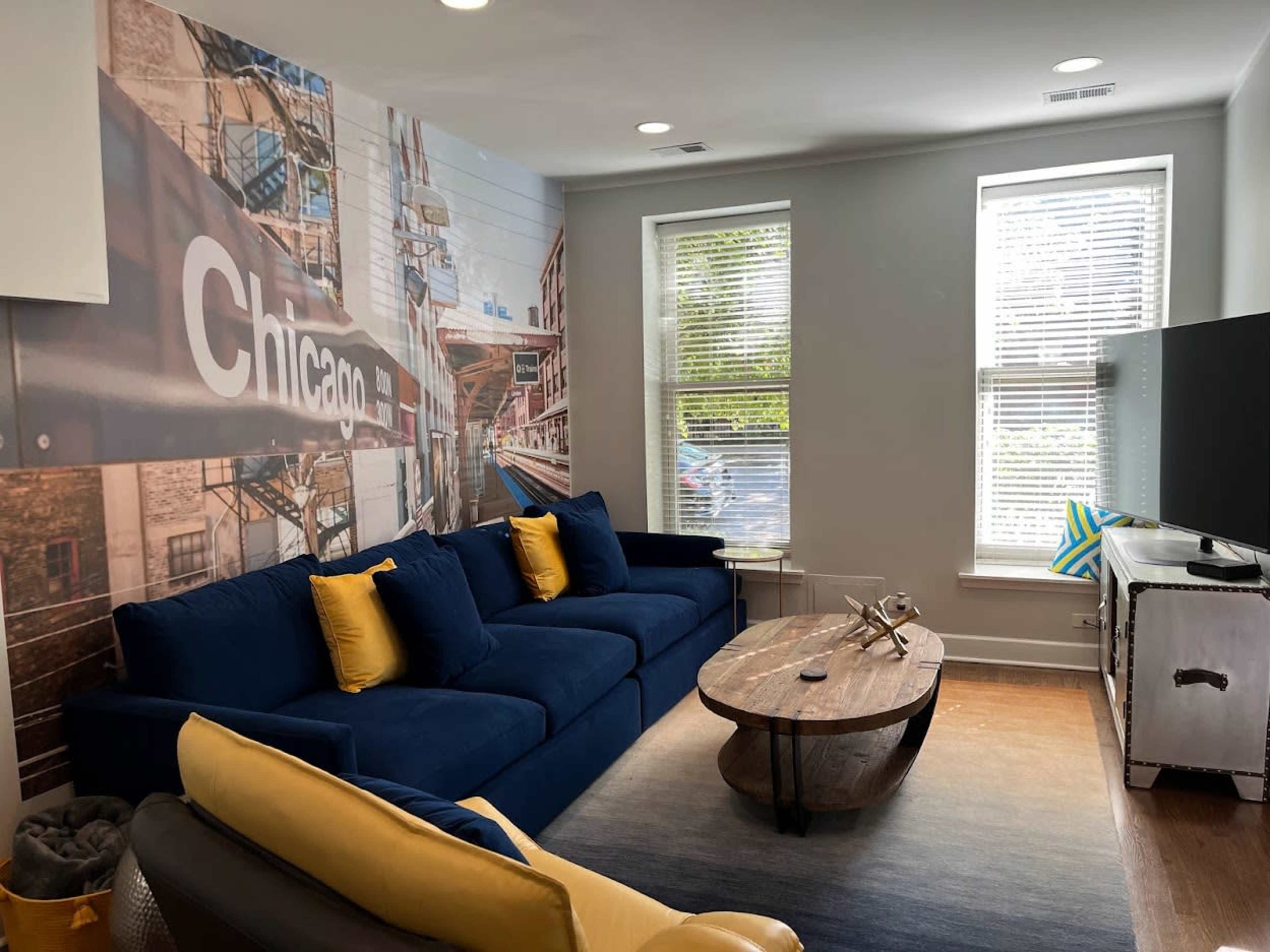 The living room features a blue couch with yellow pillows, a wooden coffee table, and a mural of Chicago on one wall, with two windows allowing natural light.