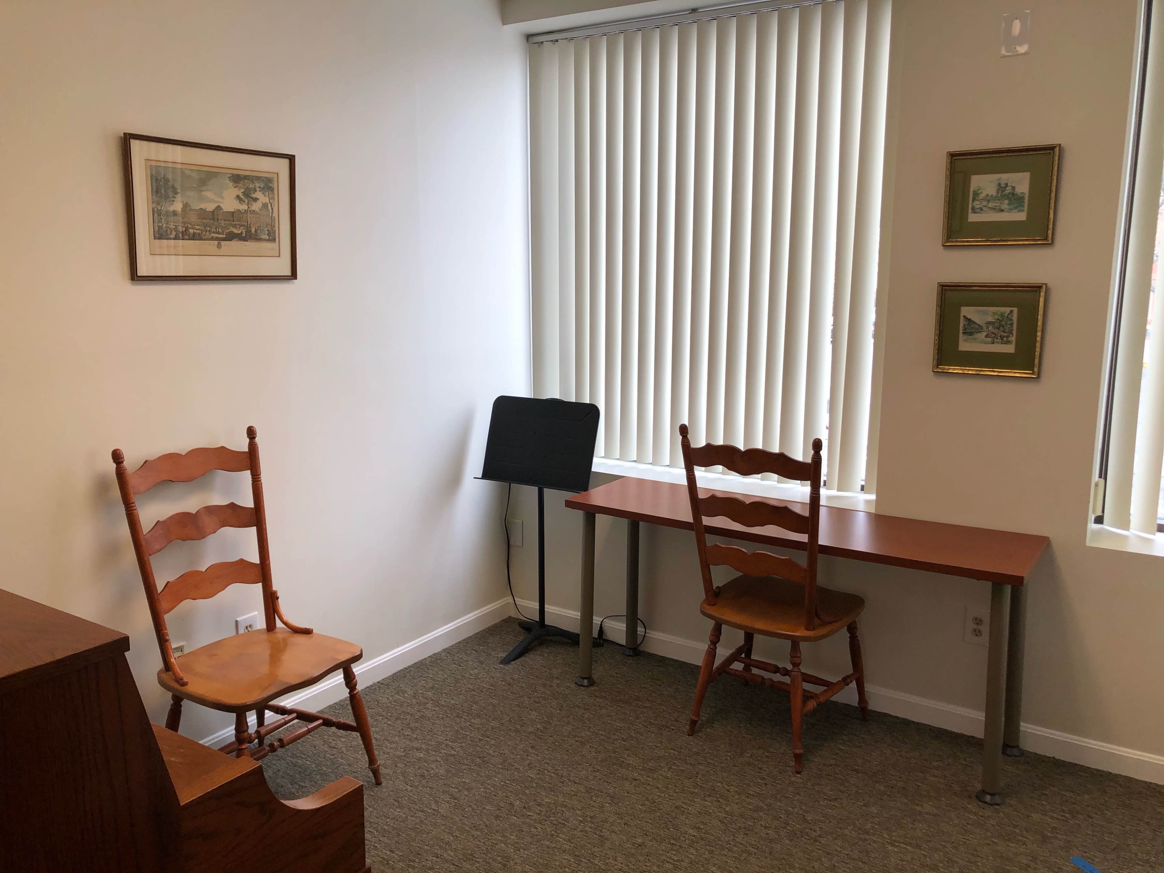A small room with two wooden chairs, a desk, a music stand, and framed pictures on the wall, illuminated by natural light from a window with vertical blinds.