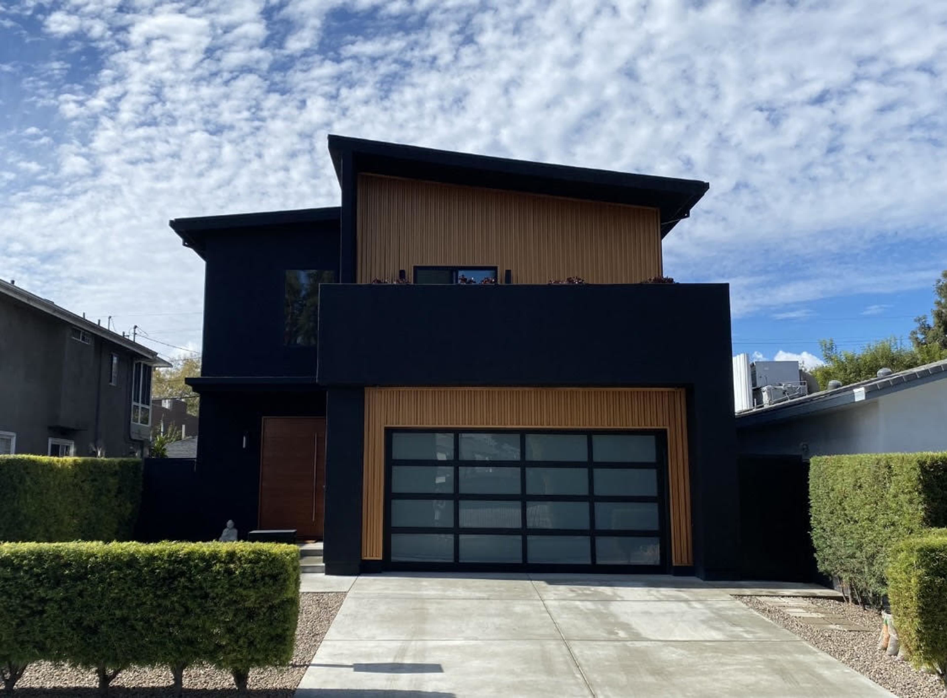 The image shows a modern two-story house with a black exterior and wood accents, featuring a large garage door and well-manicured hedges in the front yard.