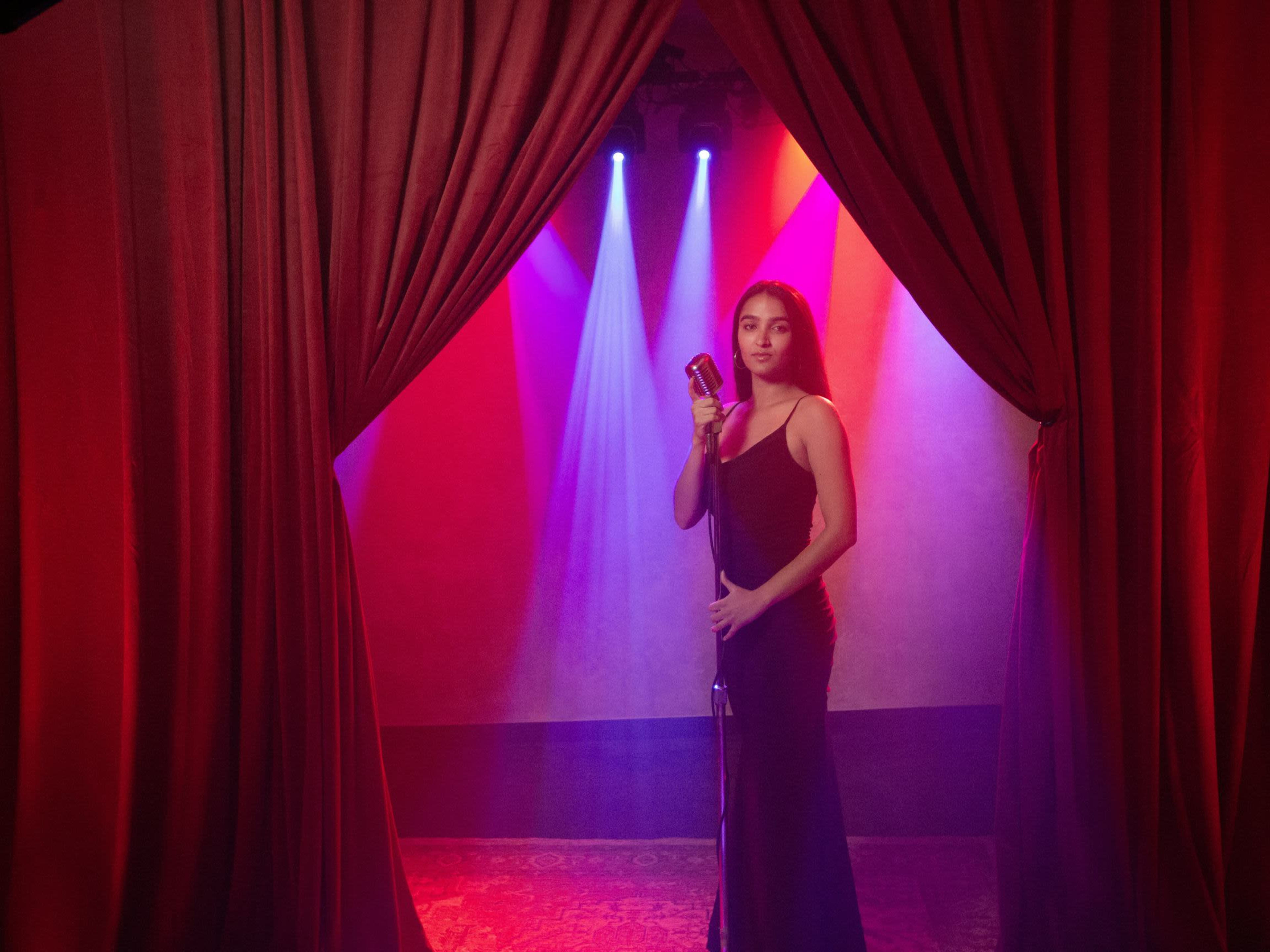 A woman stands in front of a vintage microphone on a stage, framed by red curtains and illuminated by colorful stage lights.