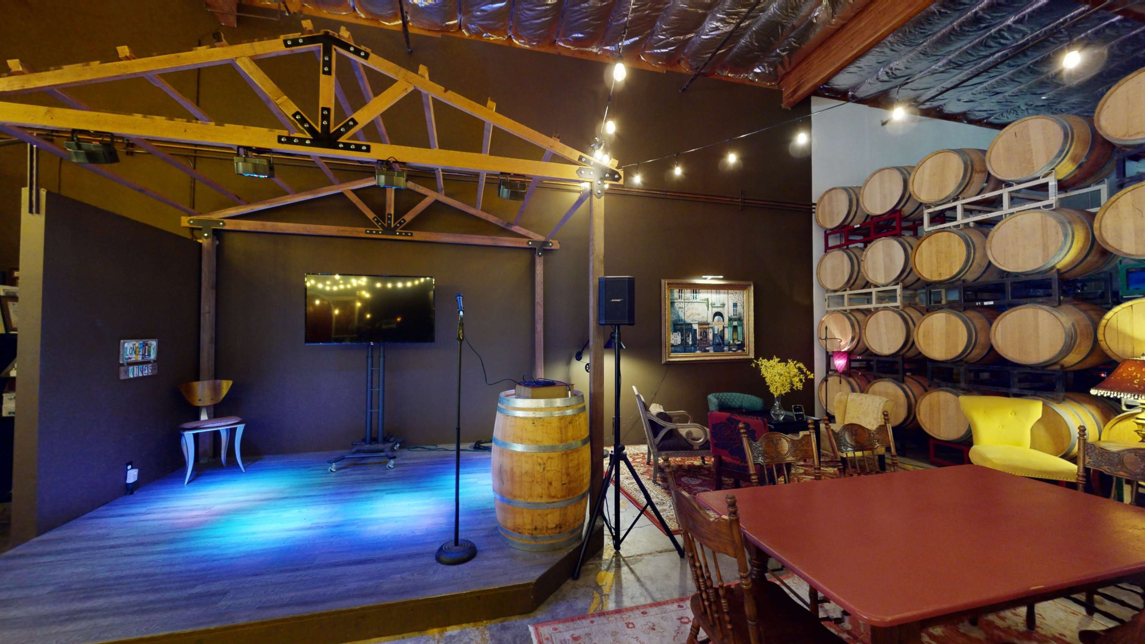 A small performance stage with a microphone and a monitor, set against a backdrop of wooden wine barrels in a cozy venue.