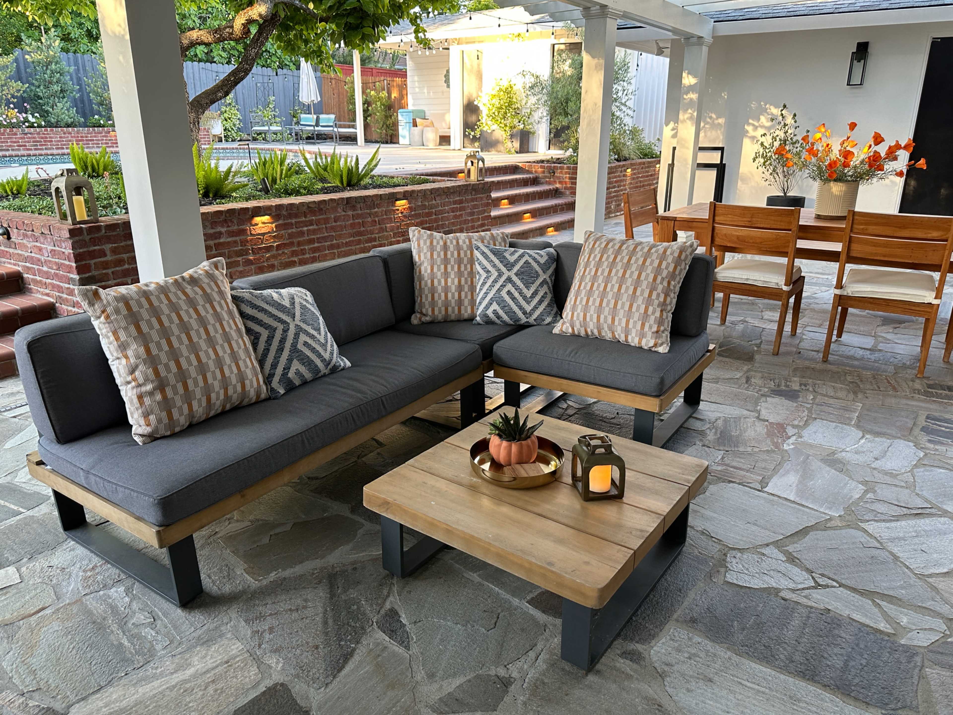 The image shows a cozy outdoor seating area featuring a gray sectional sofa with decorative pillows, a wooden coffee table with a small plant and candle, and a dining table with wooden chairs under a covered patio.