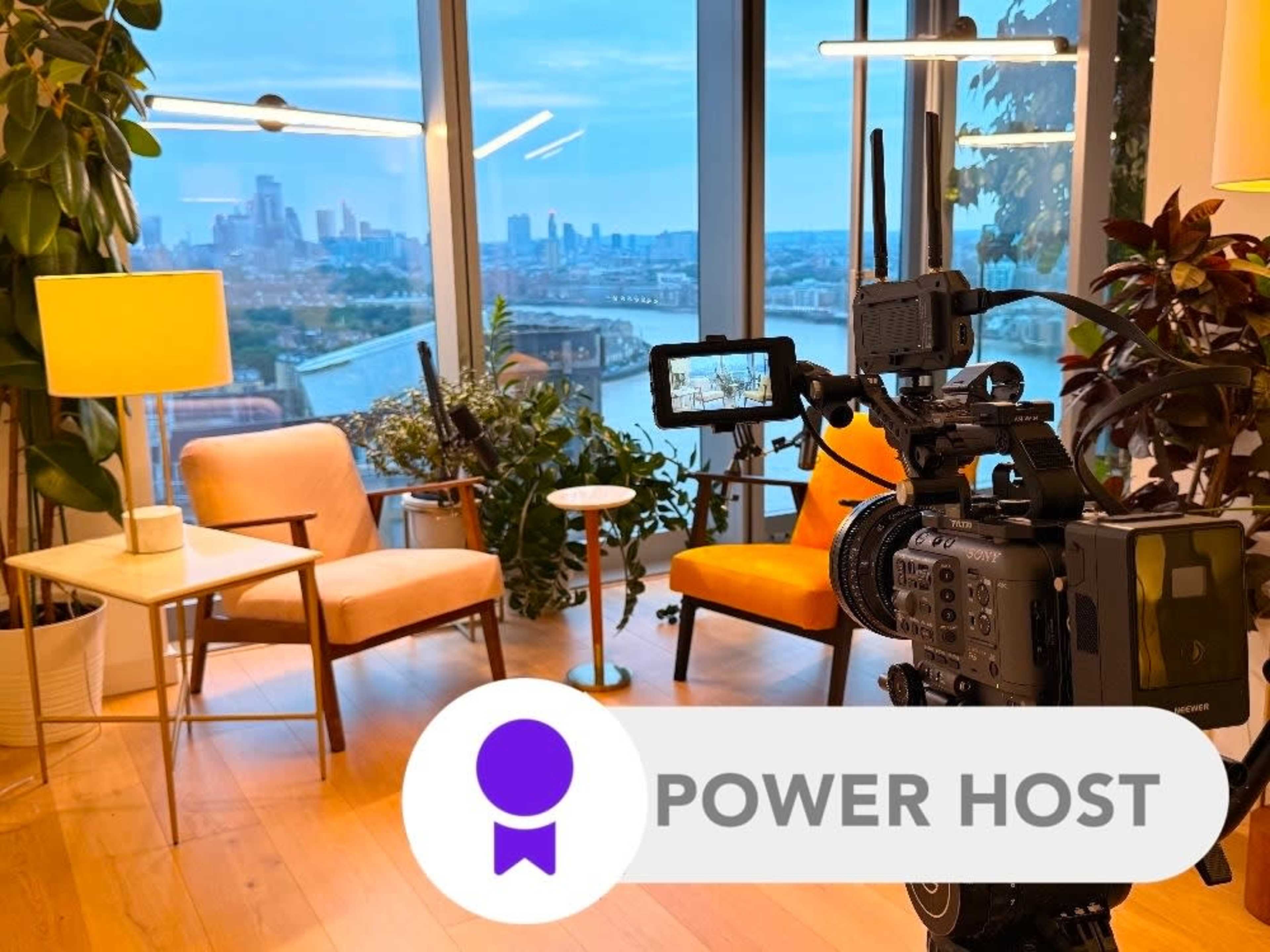 A video camera is positioned in front of two chairs and a lamp, overlooking a city skyline through a large window.