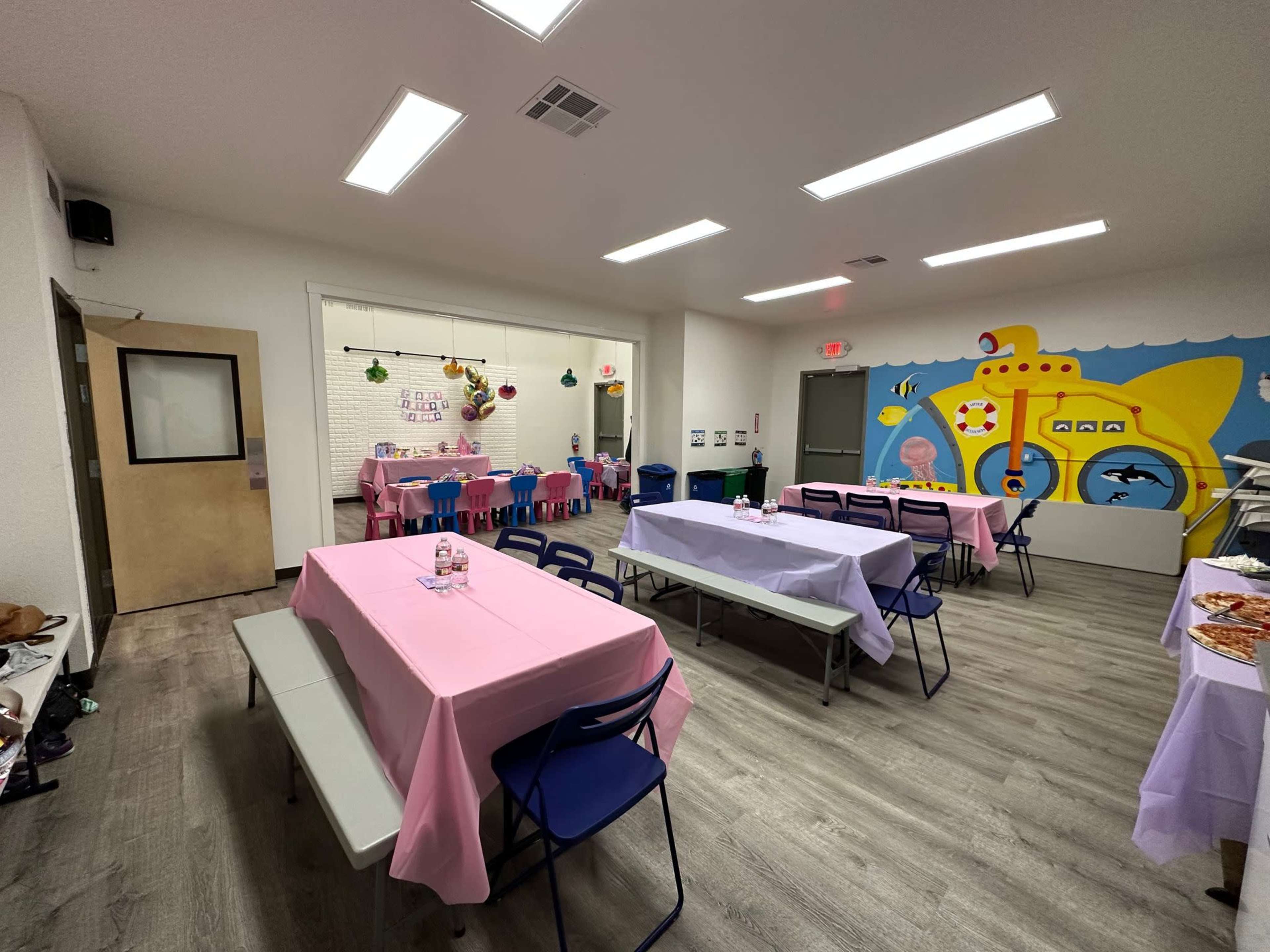 A brightly lit room with pink tables and blue chairs, decorated with a mural of a submarine and other playful elements, set up for a gathering or party.