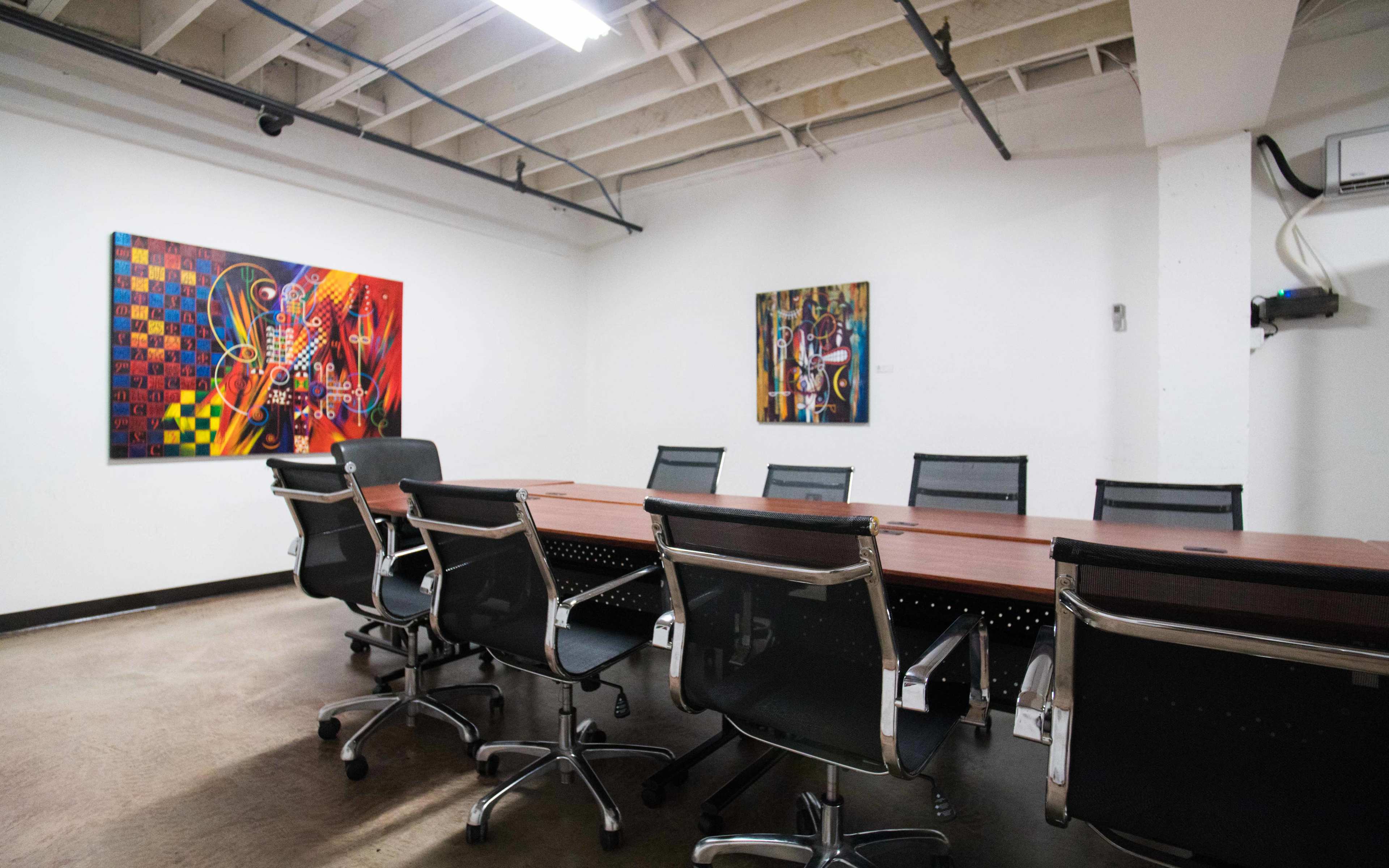 A boardroom features a long wooden table surrounded by black chairs, with colorful artworks hanging on the white walls.