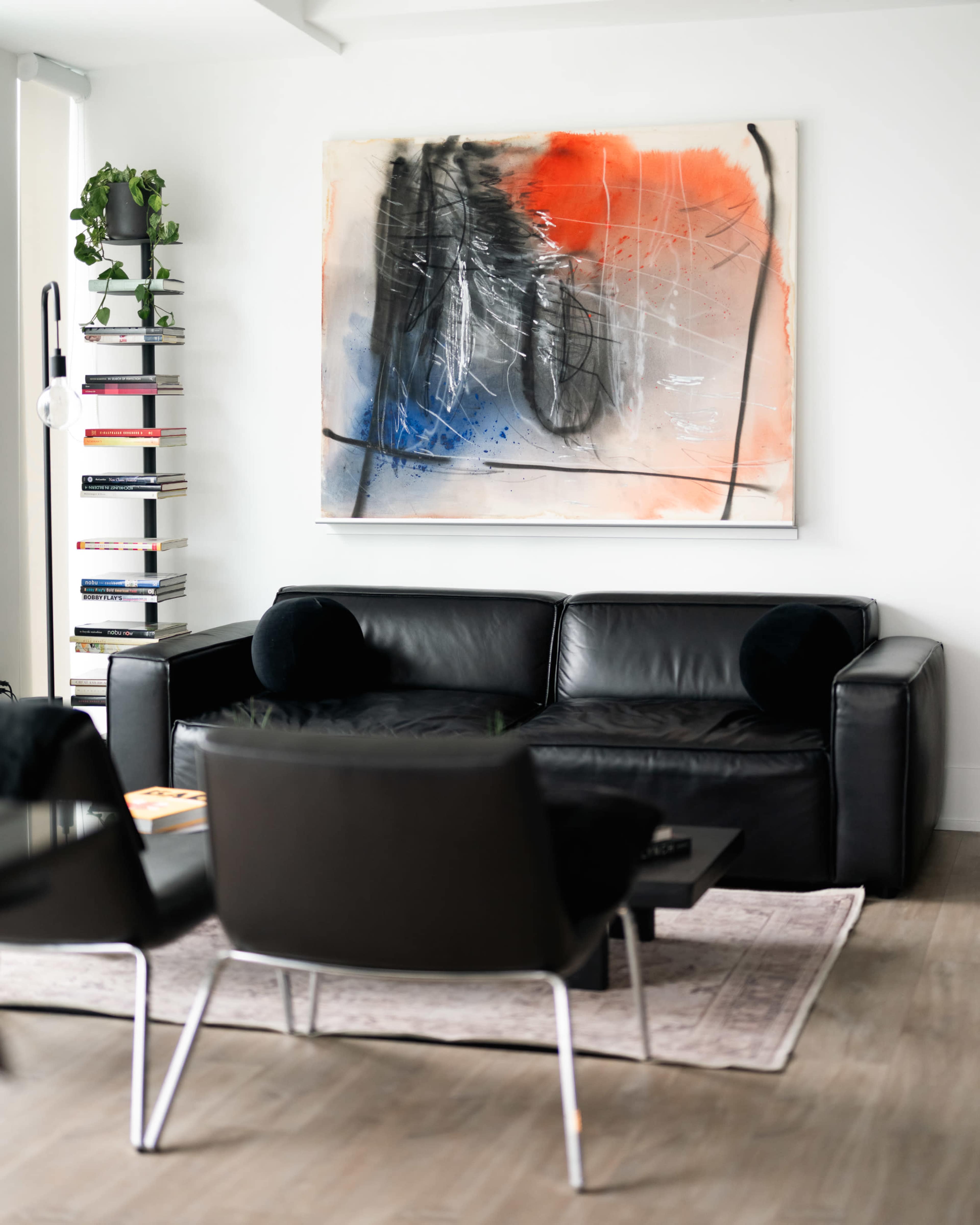A black leather sofa is positioned across from two modern chairs, with a large abstract painting hanging on the wall and a bookshelf filled with books nearby.