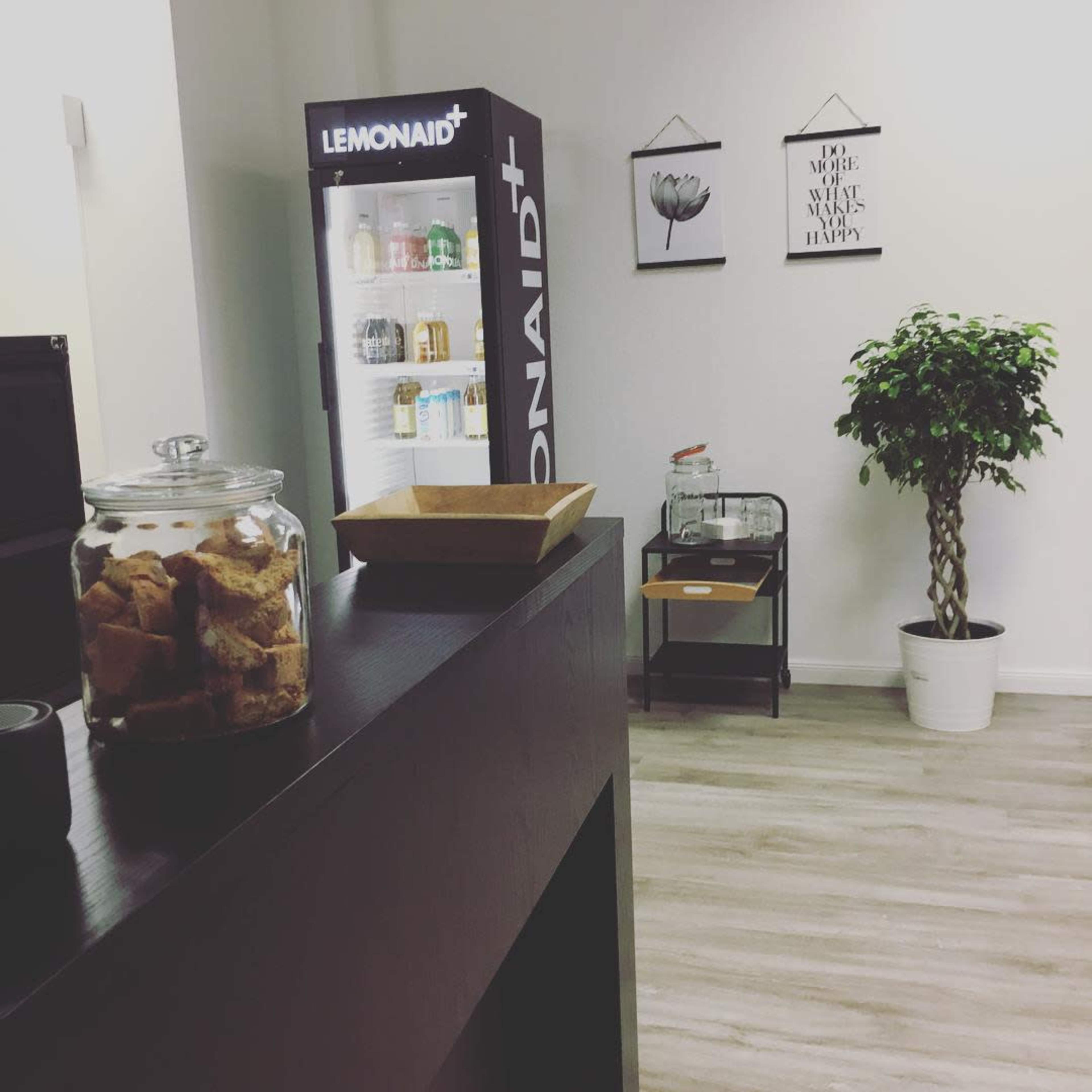 The image shows a bright, modern room with a glass-fronted refrigerator, a plant in a white pot, and a table displaying snacks.