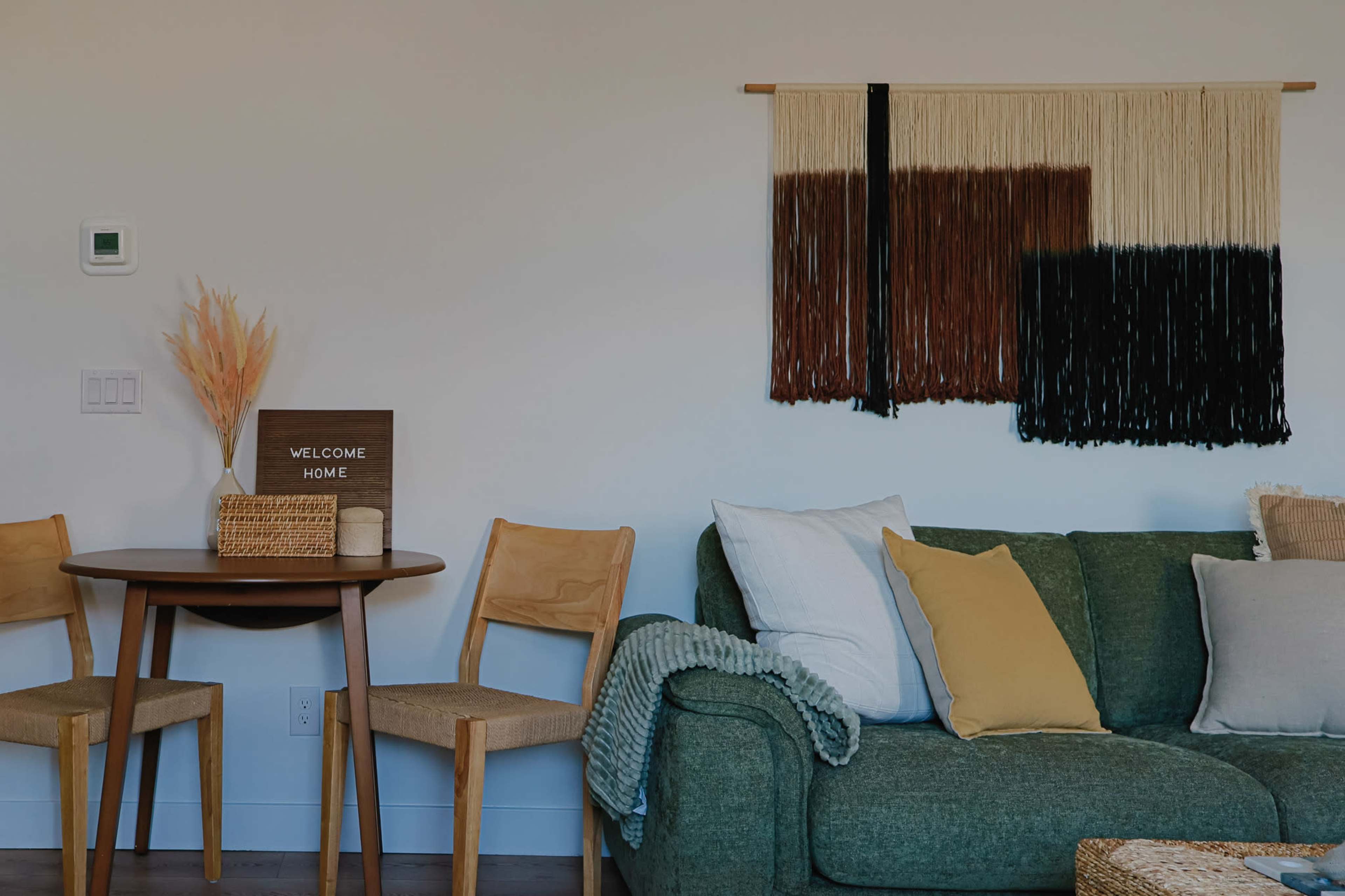 The image shows a cozy living space featuring a green sofa with various pillows, a round dining table with two chairs, and a macramé wall hanging above.