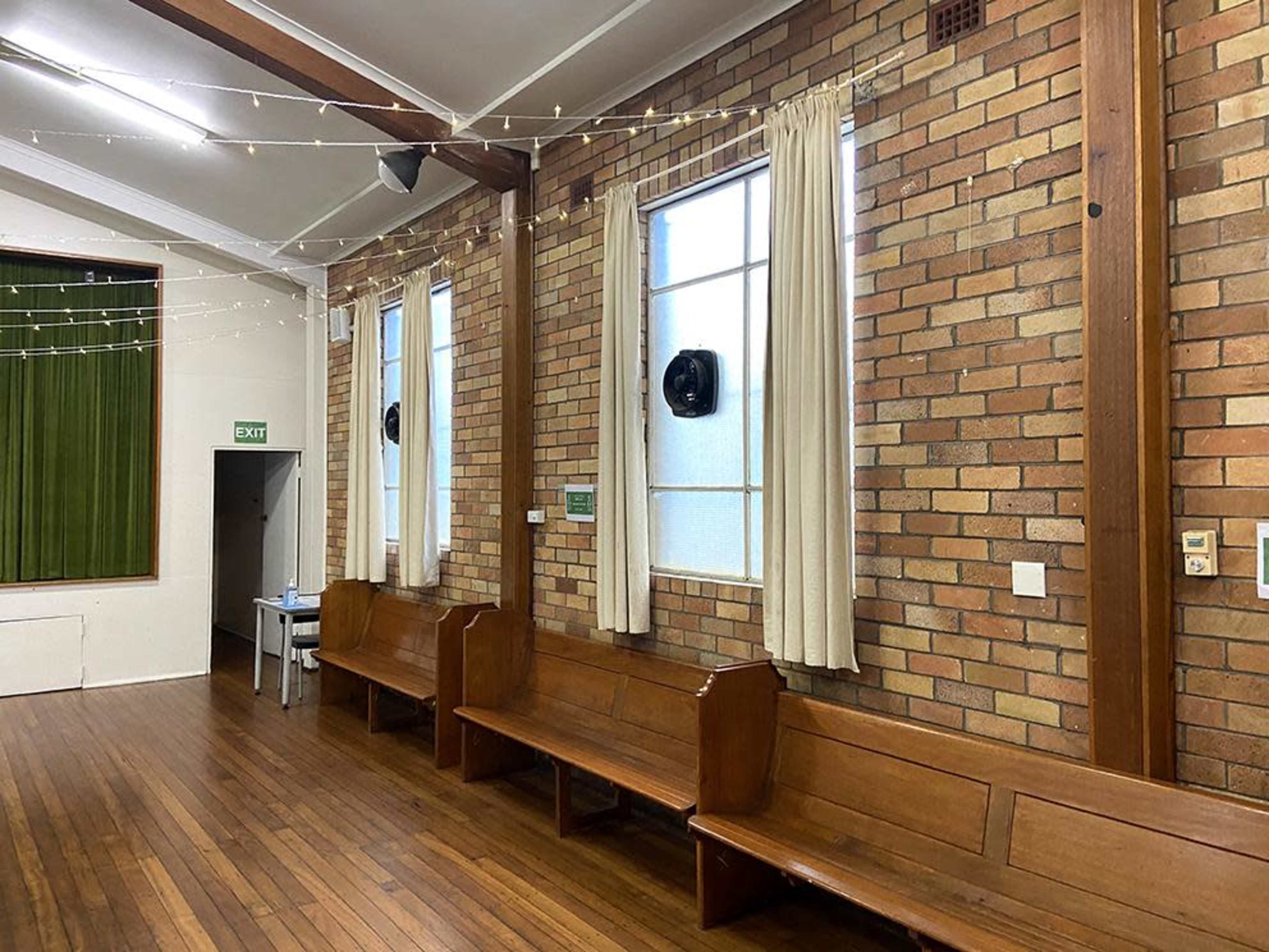 The image shows a wooden-floored hall with brick walls, featuring several long wooden benches along the sides and a curtain-covered window.