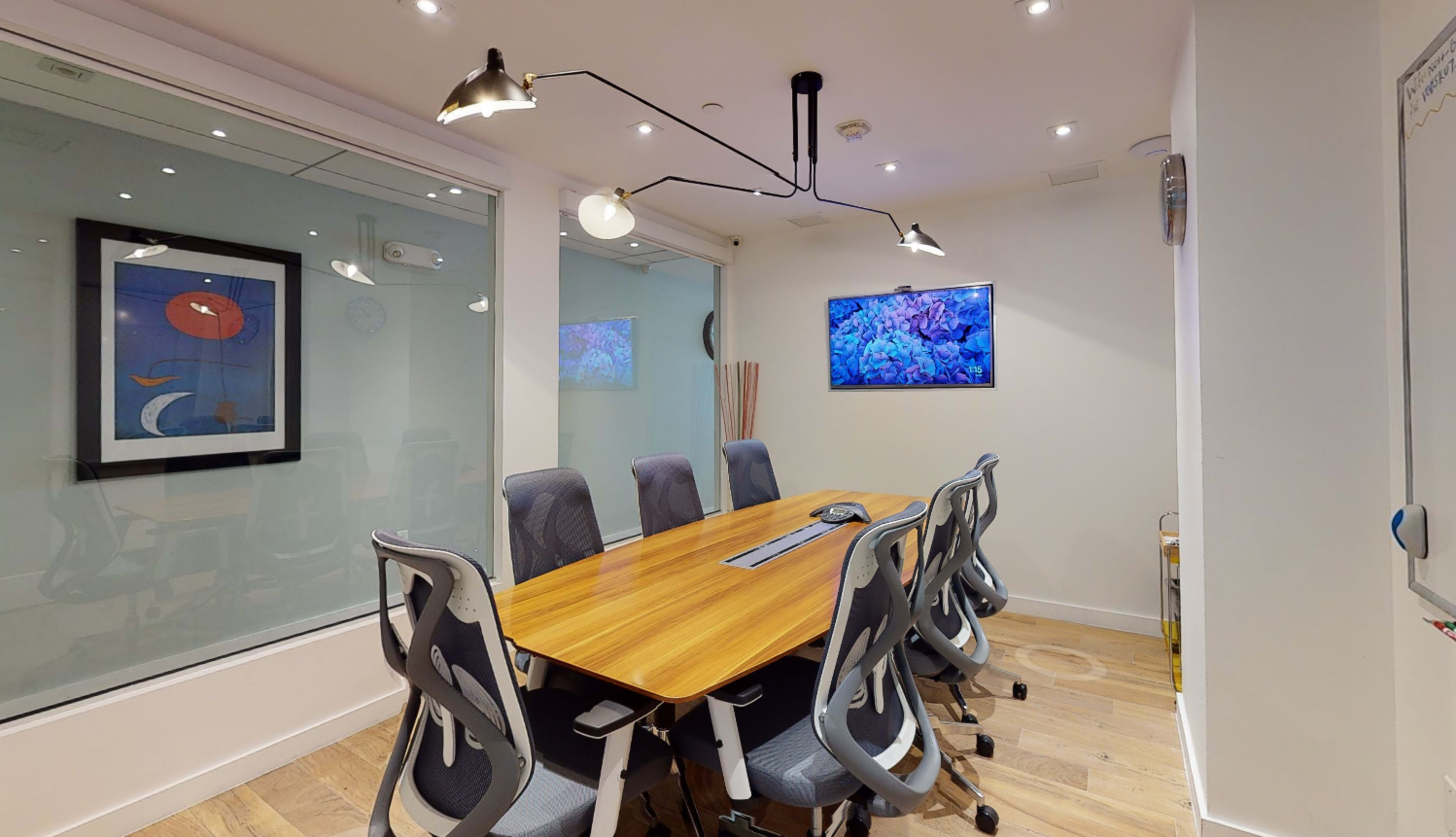 A modern conference room features a long wooden table surrounded by ergonomic chairs, with large glass windows, artistic wall decor, and a digital screen displaying an image.
