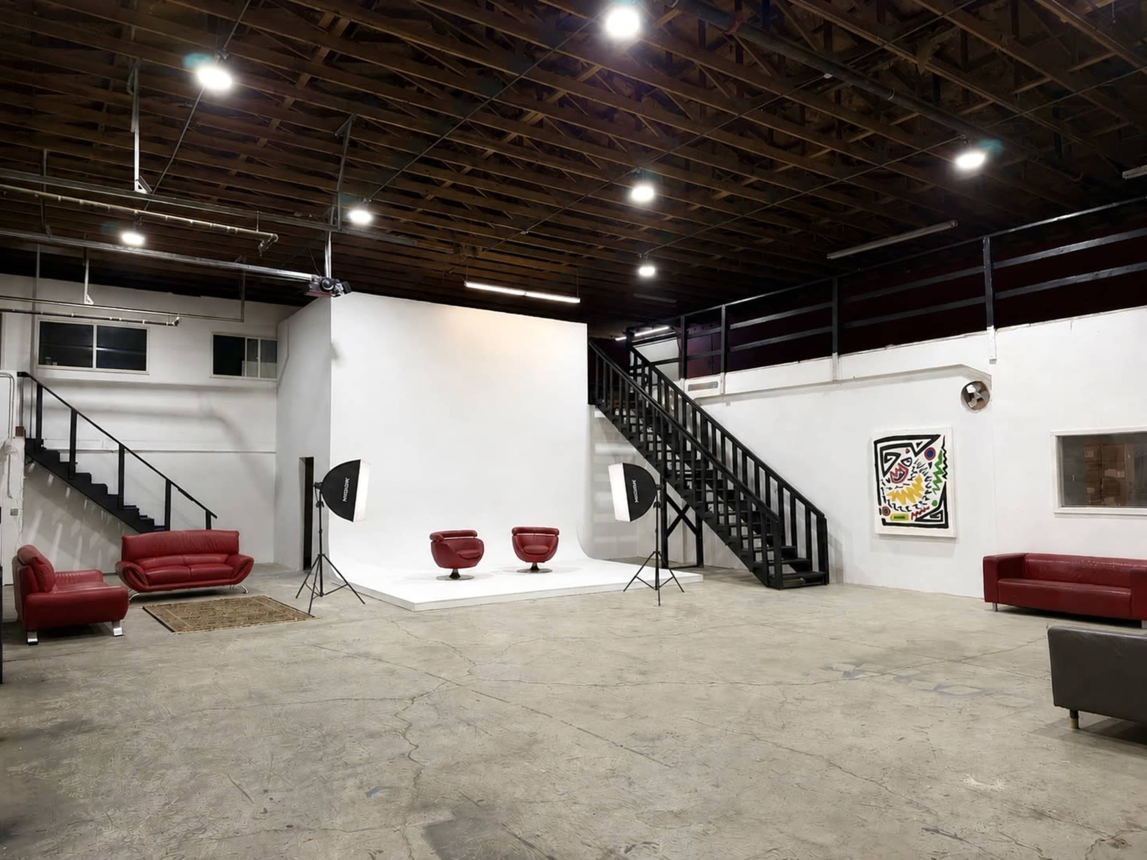 The image shows a spacious studio with a white backdrop, two red chairs, and a staircase leading to a second level.