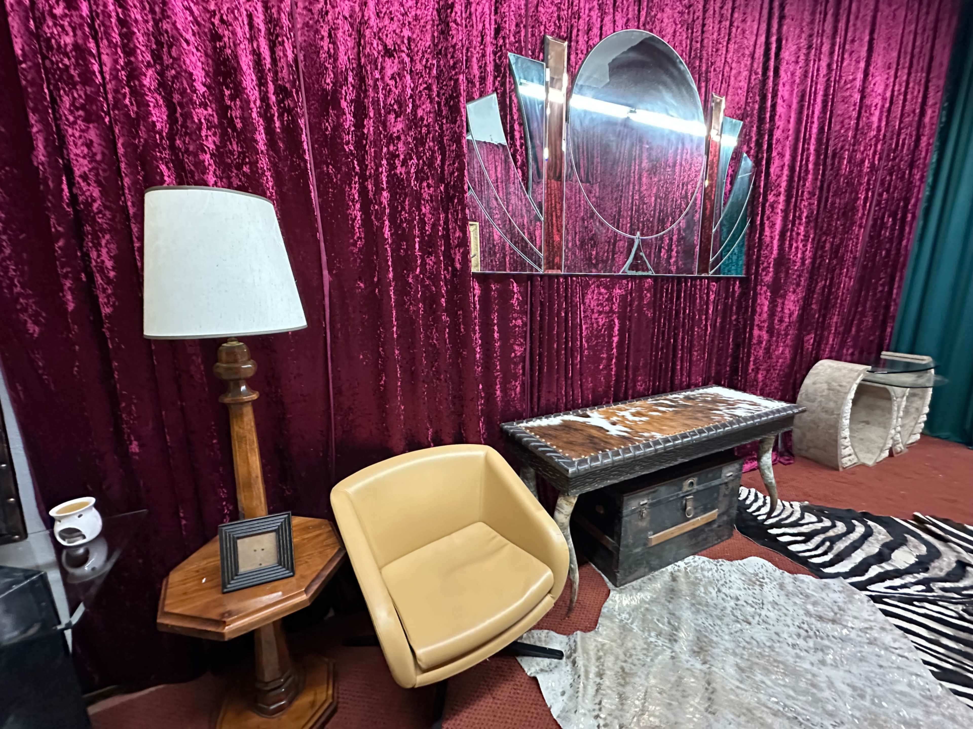 The scene features a vintage-inspired room with a plush burgundy curtain backdrop, a round mirror, a wooden table, an armchair, and various decorative elements.