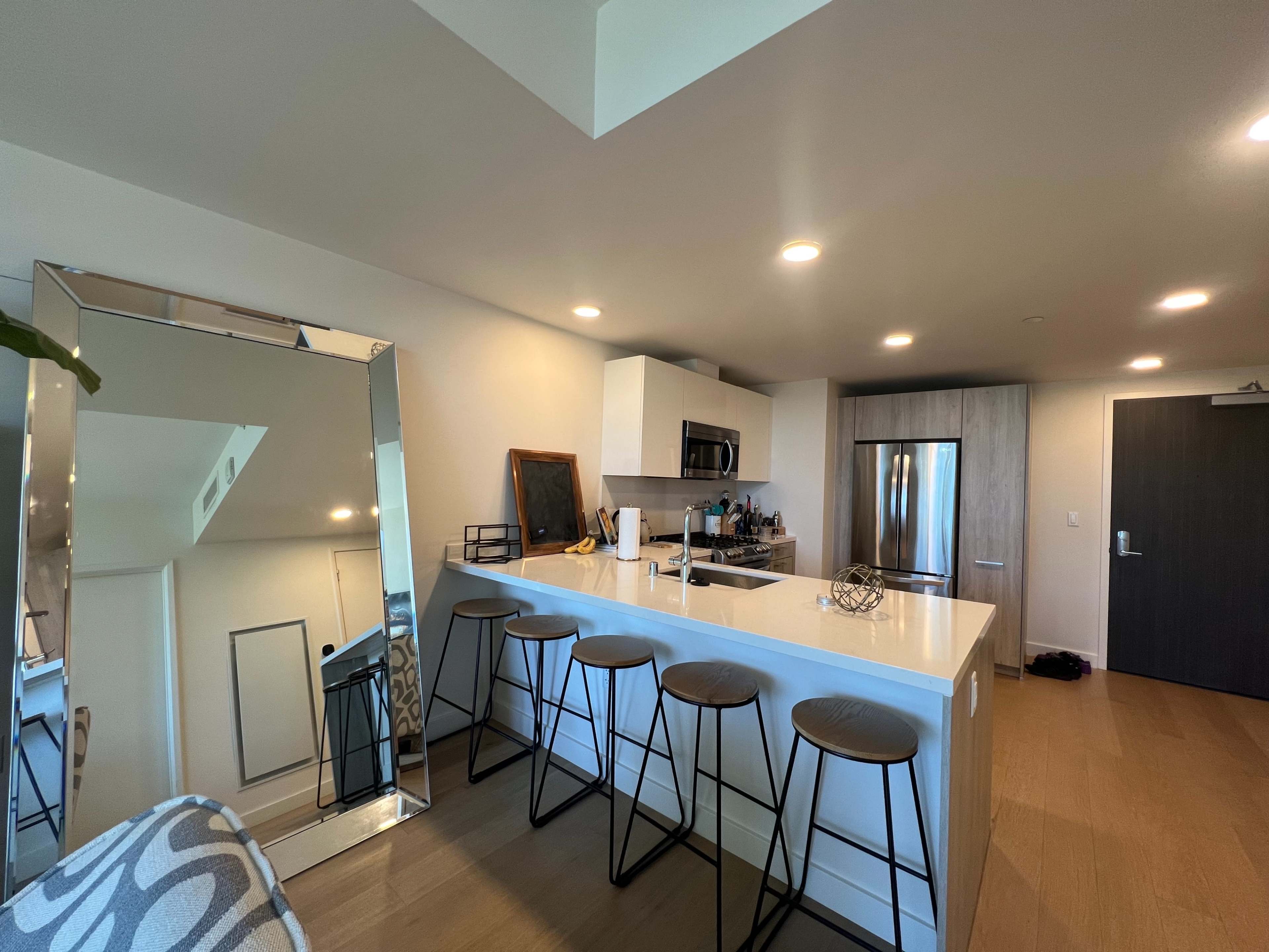The image shows a modern kitchen with a countertop, four stools, and stainless steel appliances, reflecting a spacious layout.