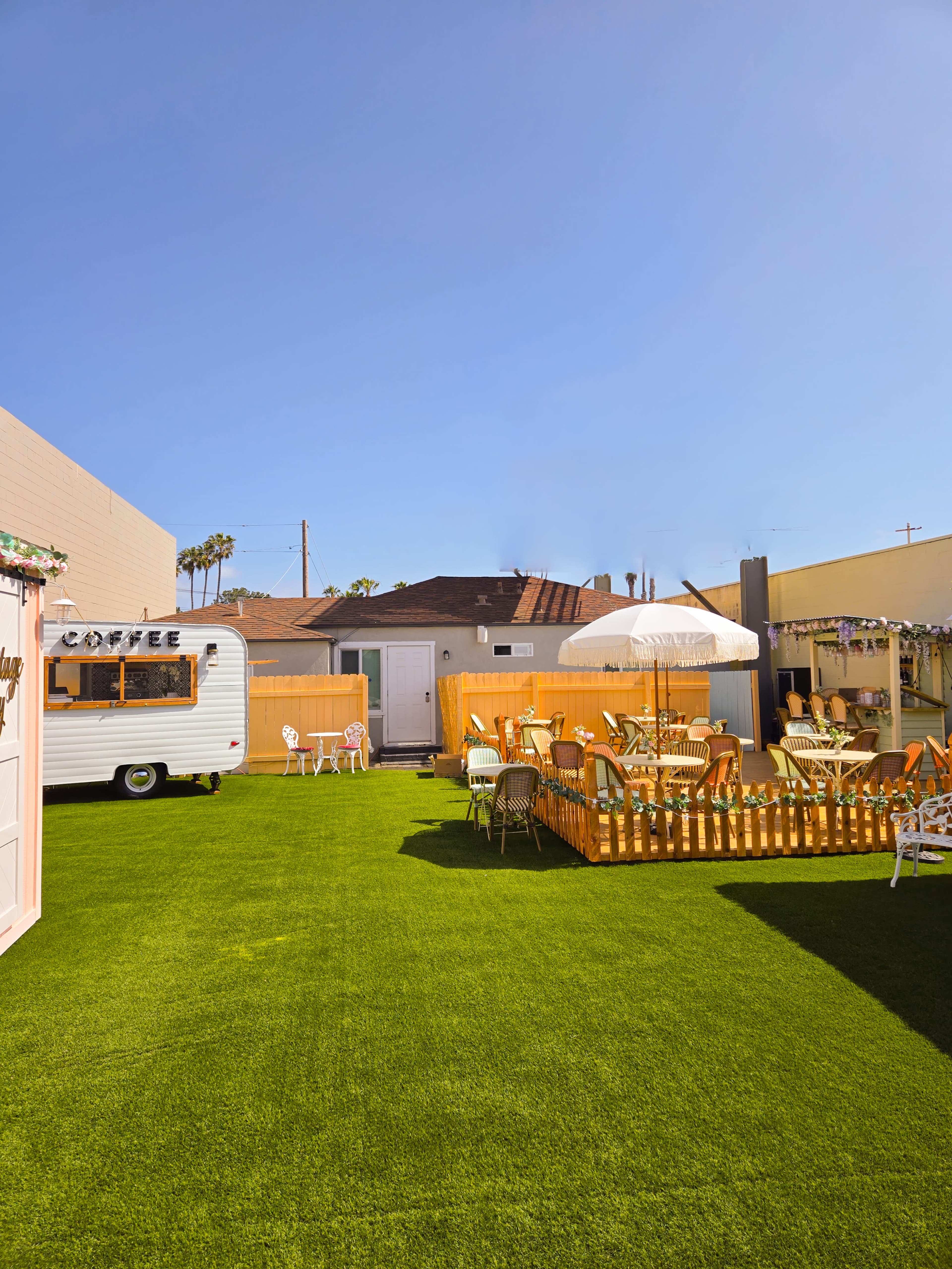 A vibrant, grassy yard with a vintage camper, outdoor seating, and a wooden fence under a clear blue sky.