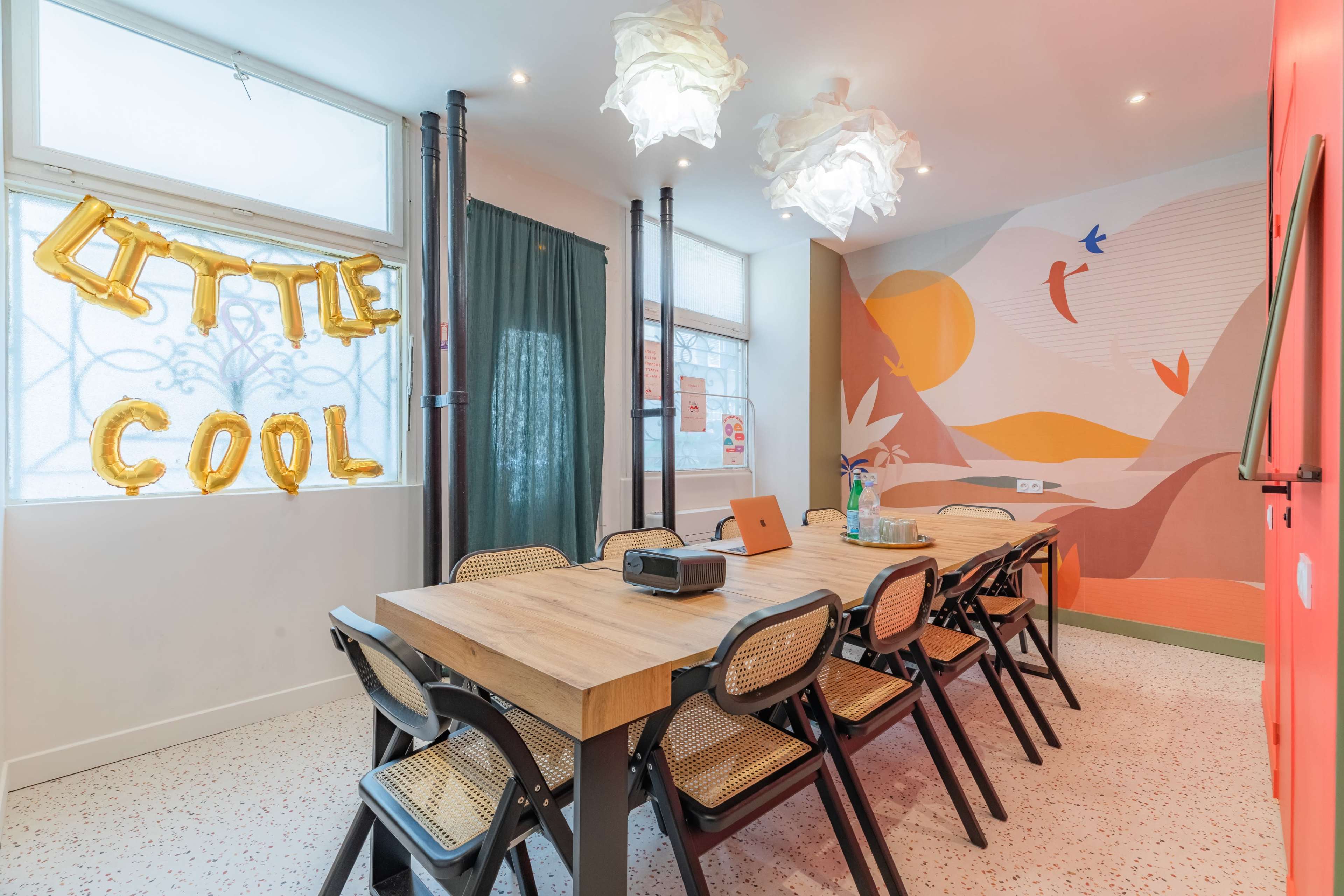 A modern meeting room with a wooden table, black folding chairs, and colorful wall art, featuring a decorative banner that reads "LITTLE COOL."