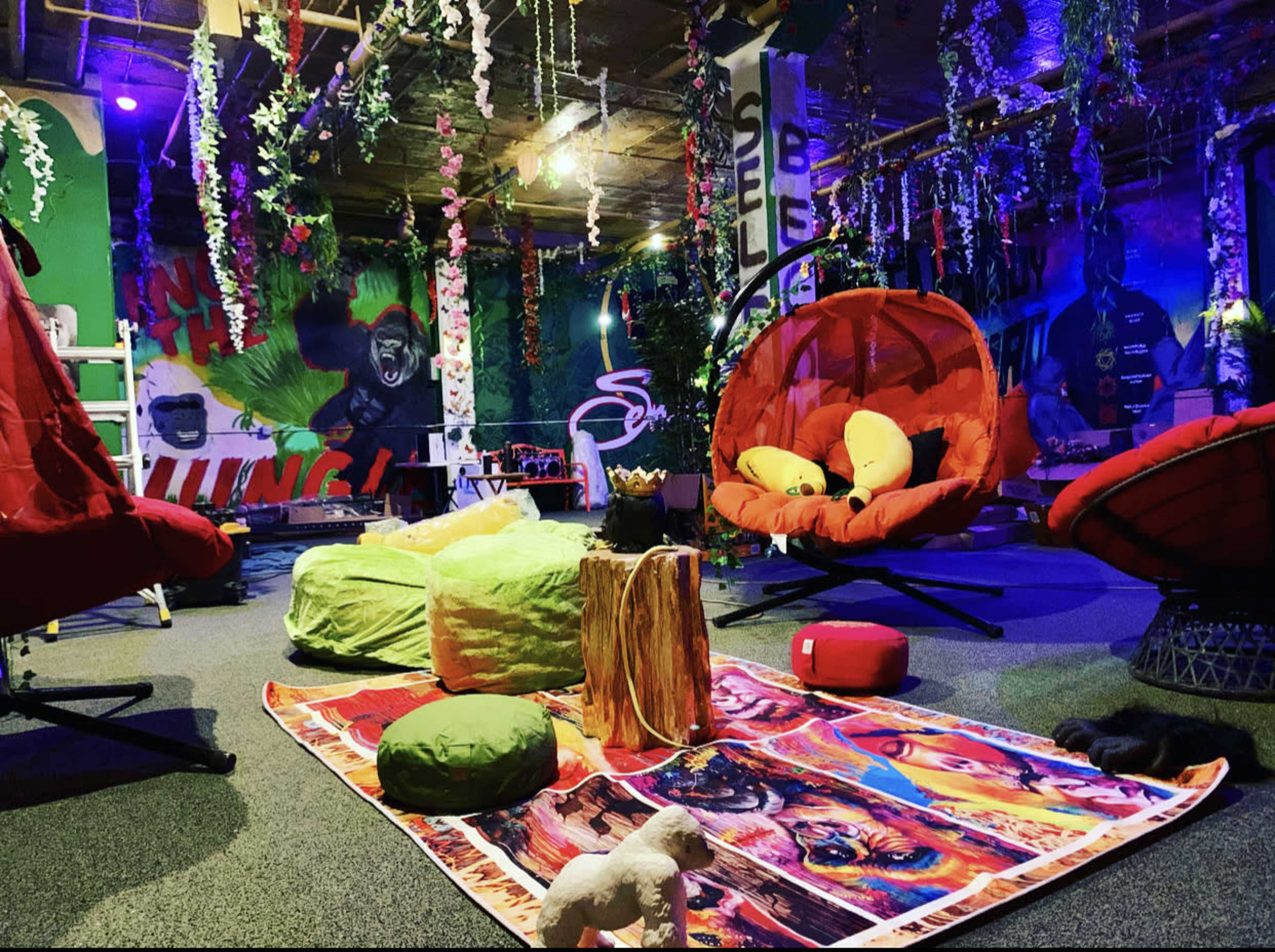 A colorful indoor lounge area with hanging foliage, vibrant murals, and various seating options, including round chairs and bean bags.