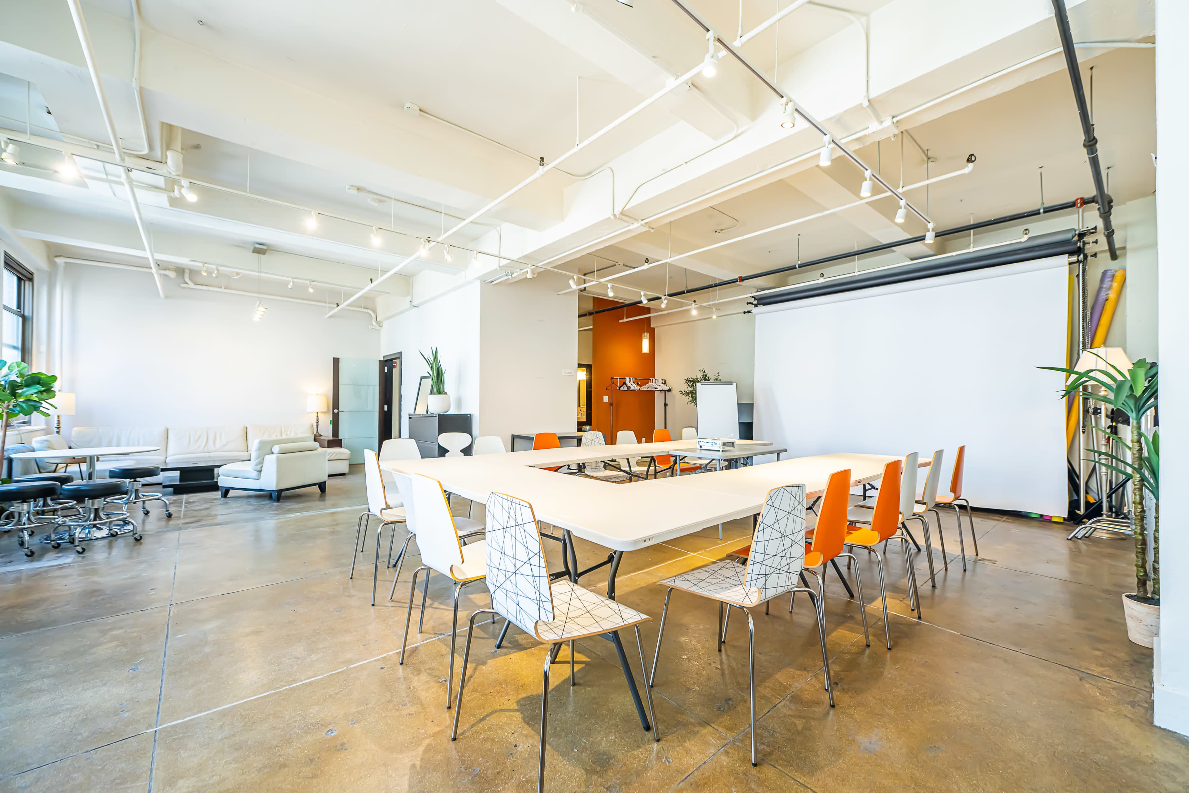 A modern meeting room features a large white table surrounded by orange and white chairs, with a projector screen and industrial-style decor.