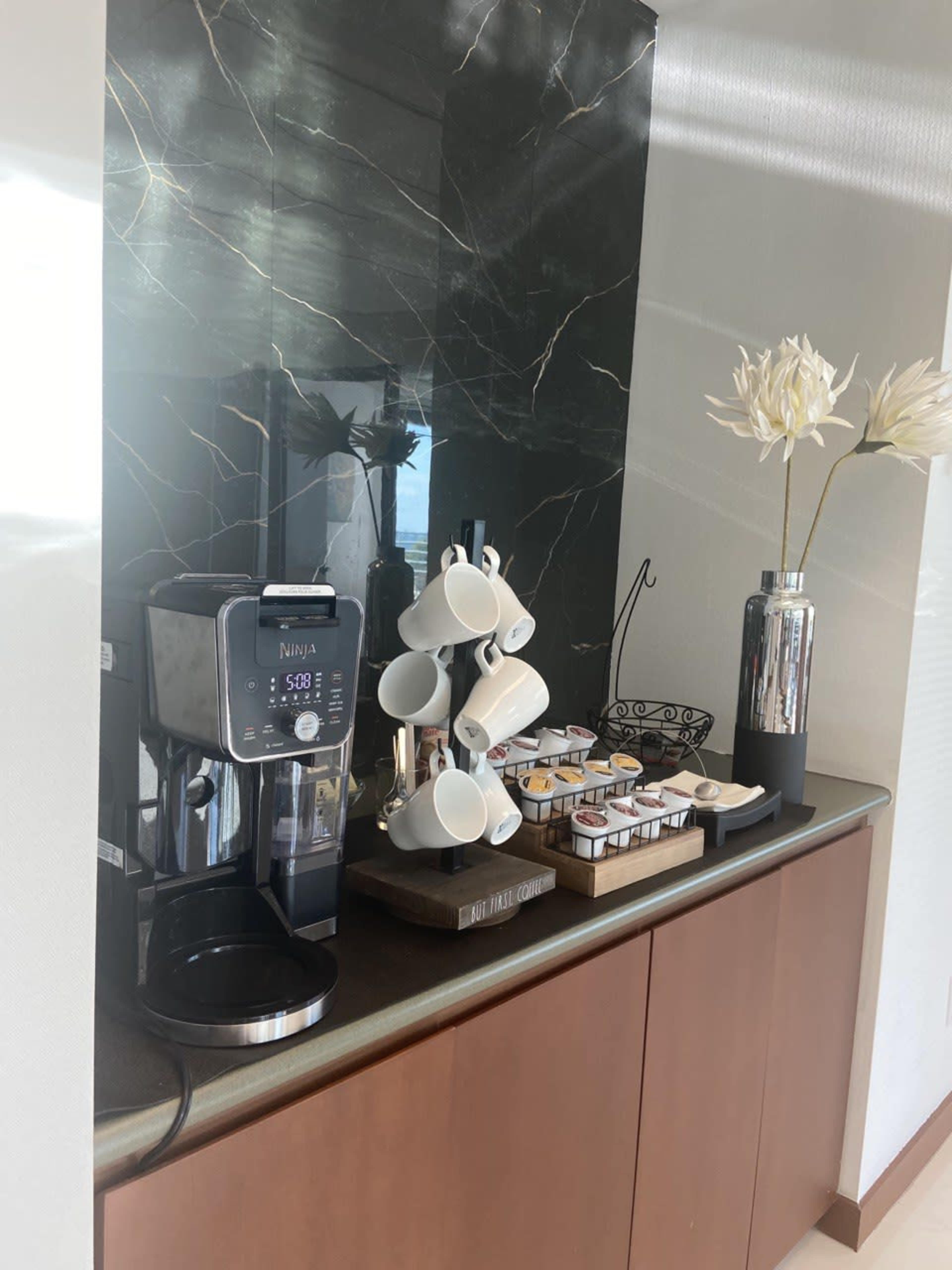 A kitchen counter with a Ninja coffee maker, a tiered rack of coffee pods, a holder for cups, a decorative vase with flowers, and a dark marble backsplash.