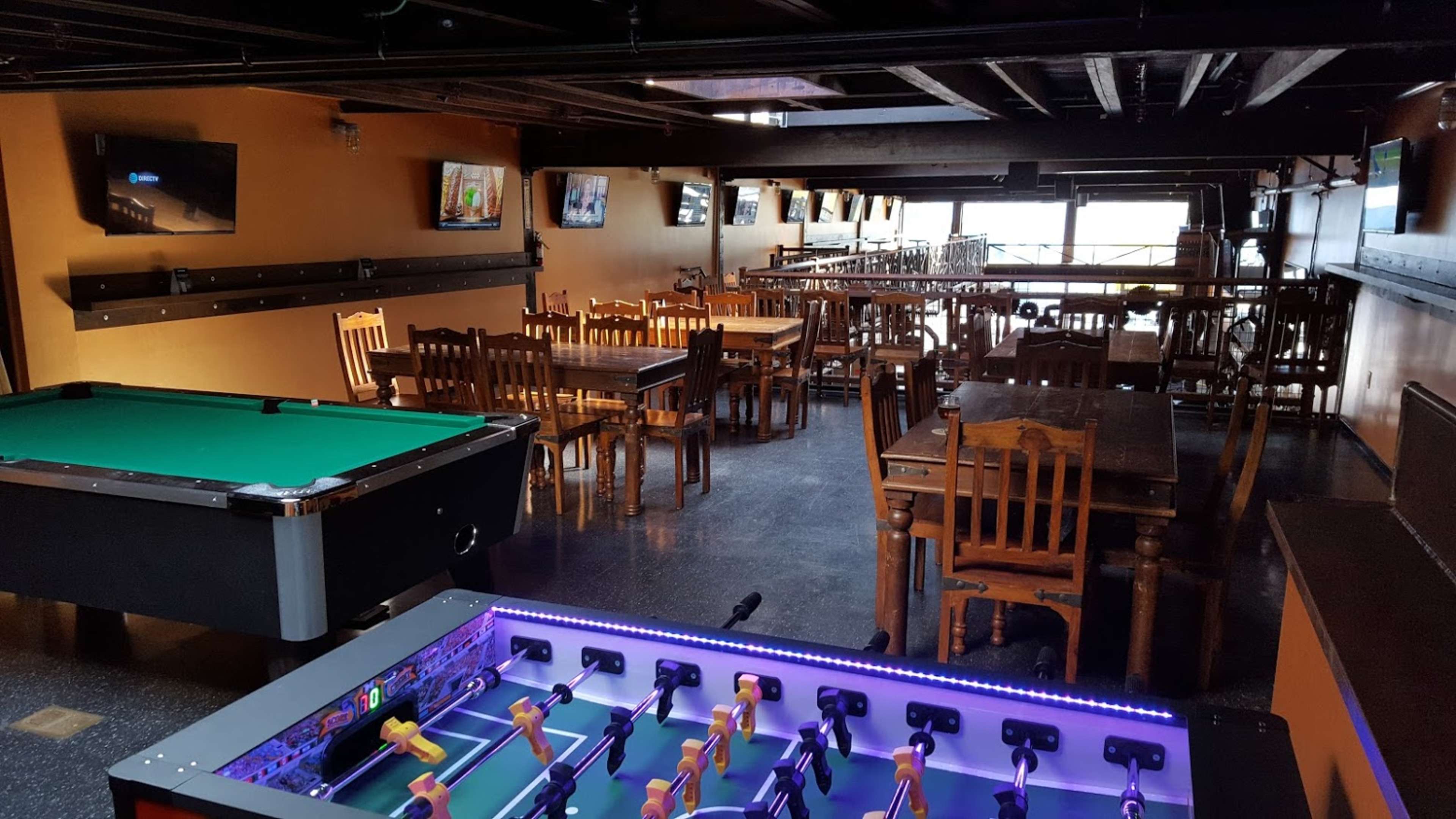 A spacious indoor venue featuring a pool table, foosball table, and several wooden dining tables with chairs.