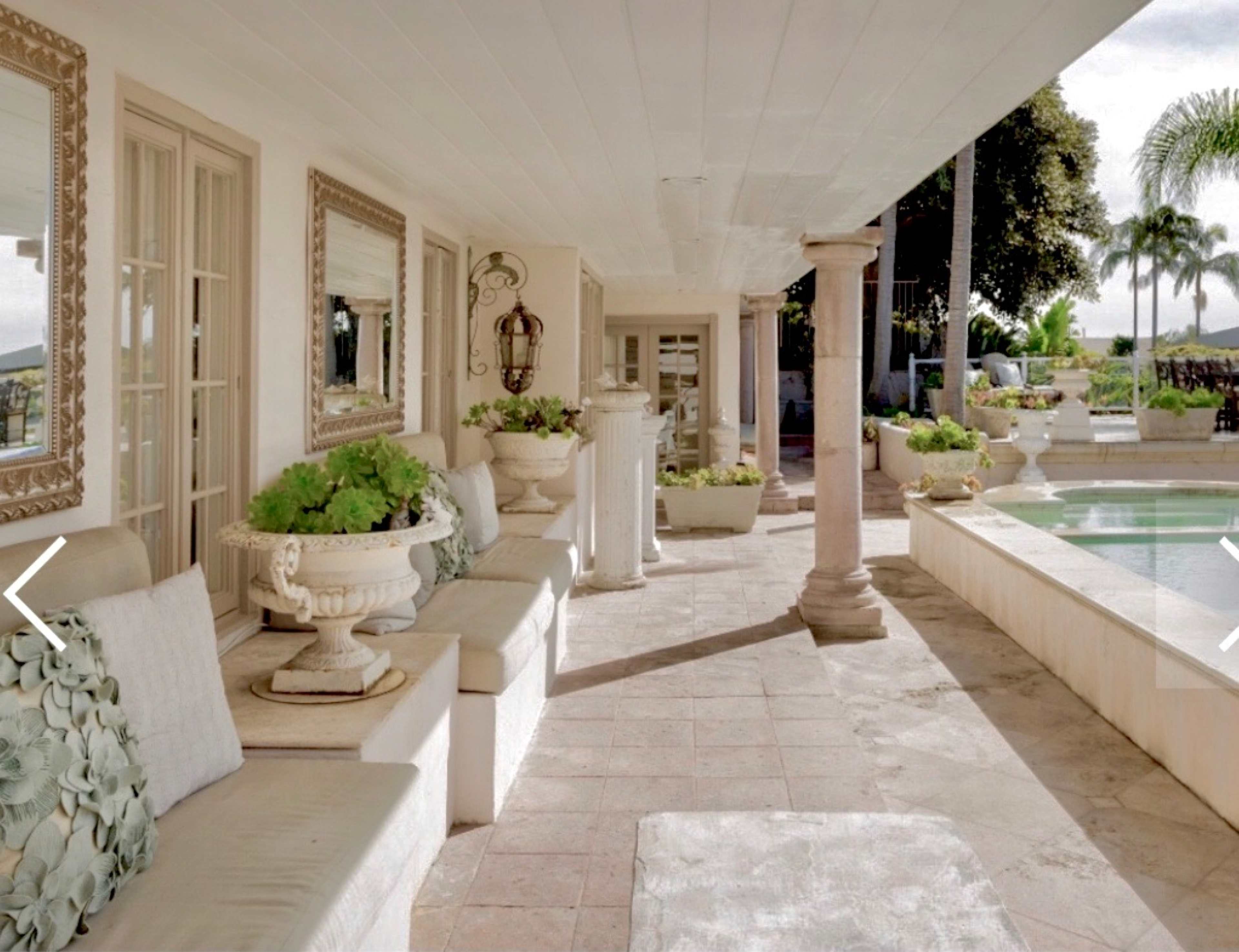 The image shows a spacious outdoor patio with seating, decorative planters, and a pool area, surrounded by greenery.