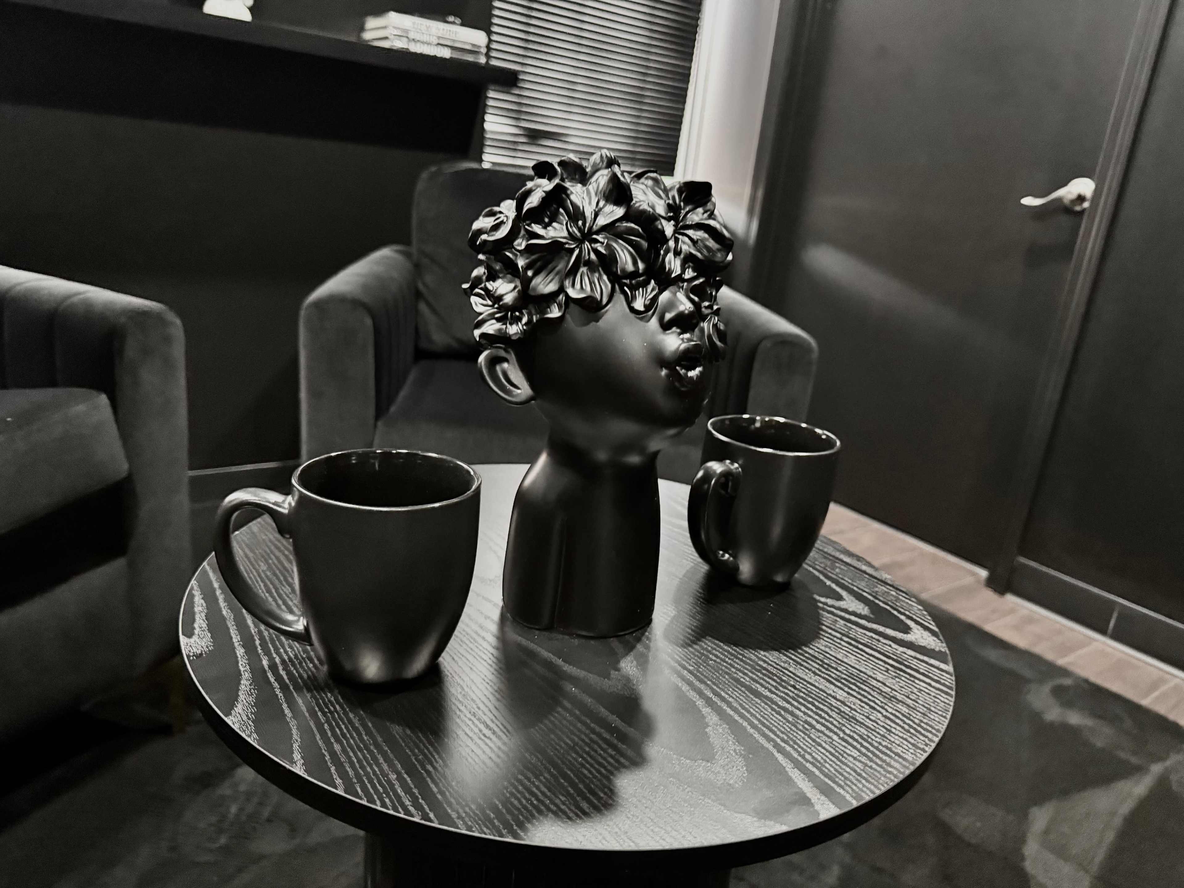 A black sculptural head with flowers on top is placed on a small table between two black coffee mugs in a dimly lit room.