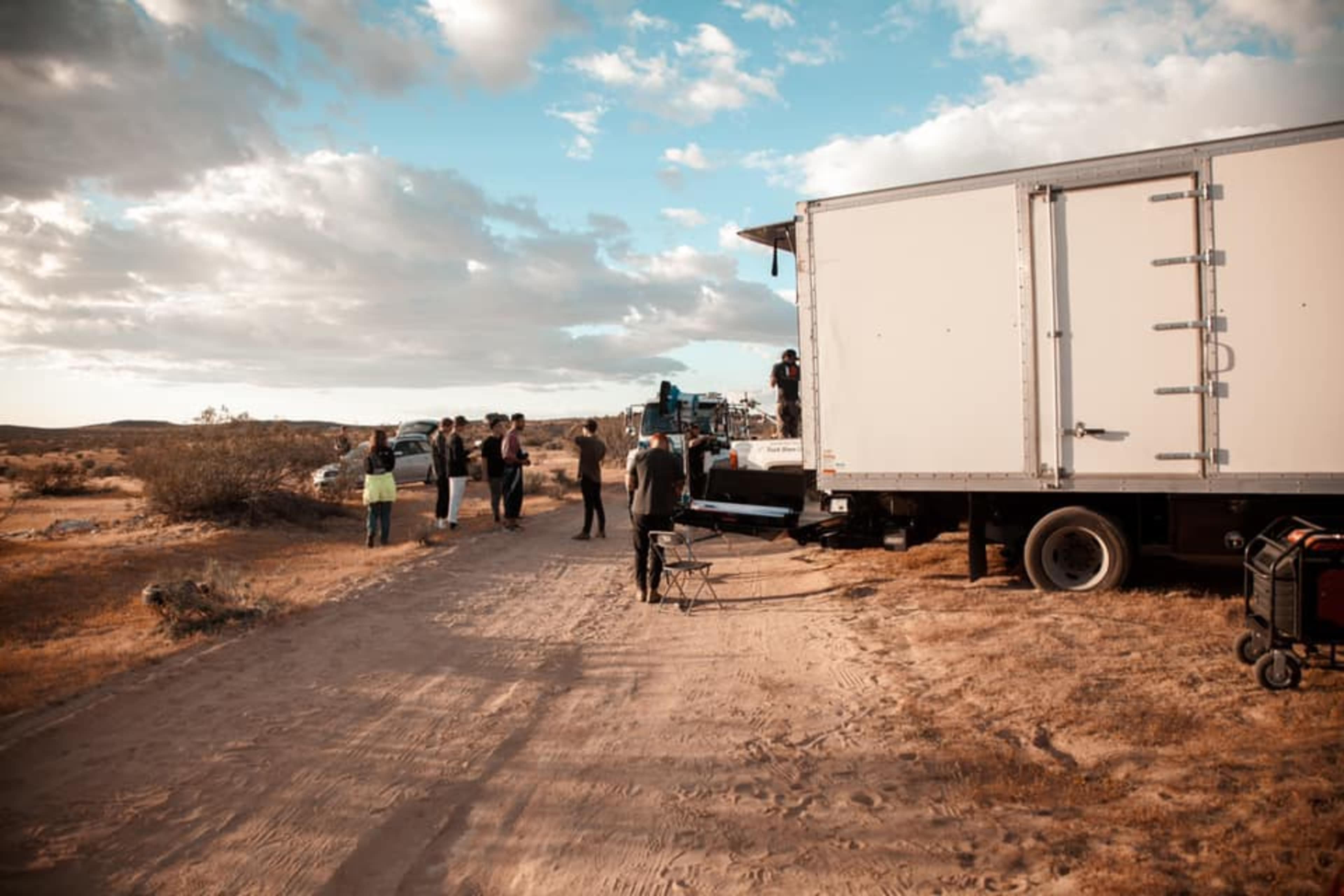 A film crew is set up near a truck on a dirt road in an open desert landscape with scattered clouds in the sky.