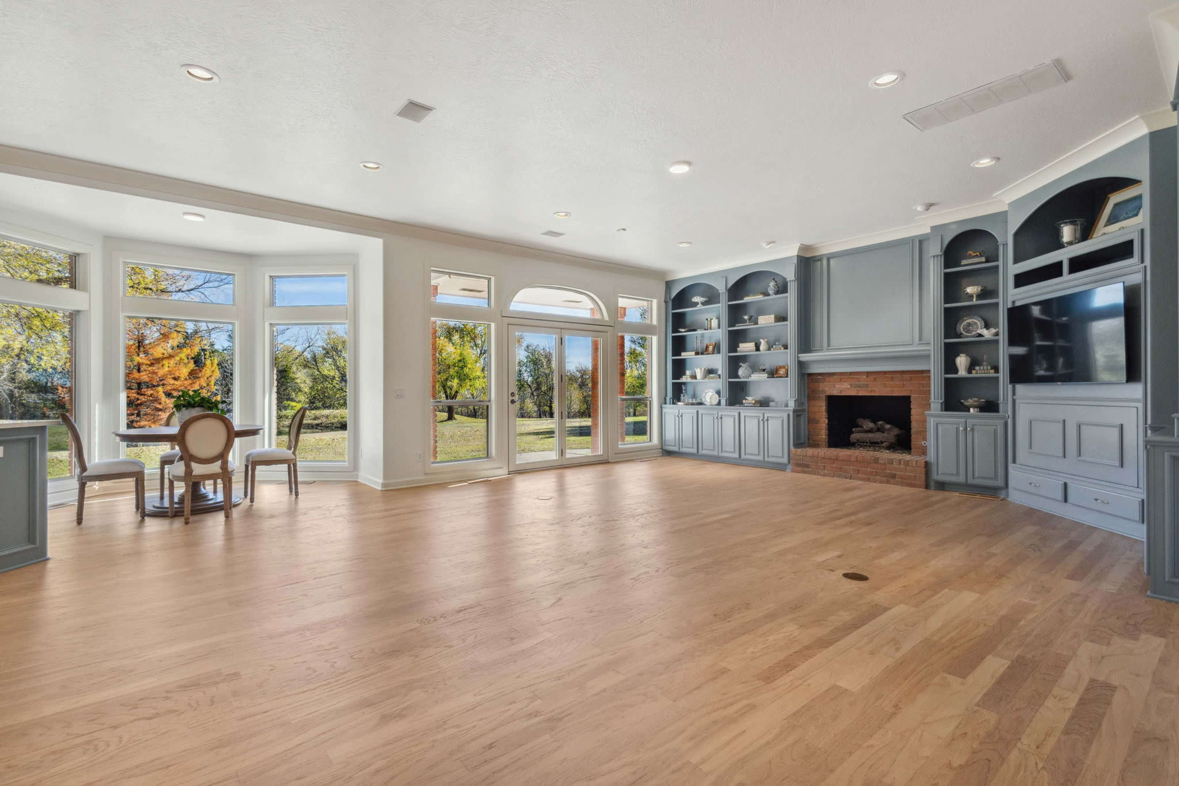 The image shows a spacious living area featuring large windows, wooden flooring, built-in shelves, and a fireplace.