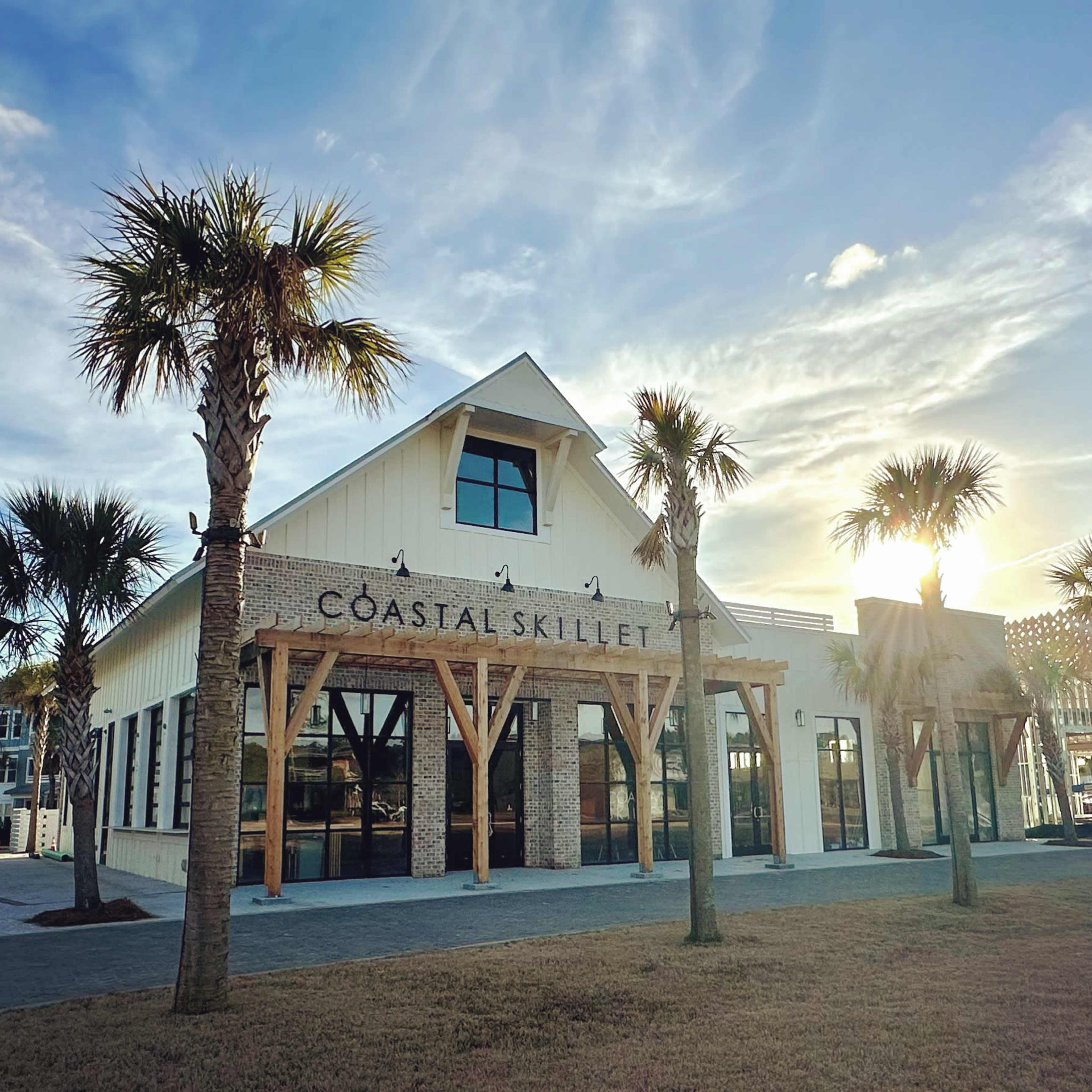 The image shows a modern white building with a wooden beam entrance and large windows, featuring the sign "Coastal Skillet," surrounded by palm trees under a bright sky.