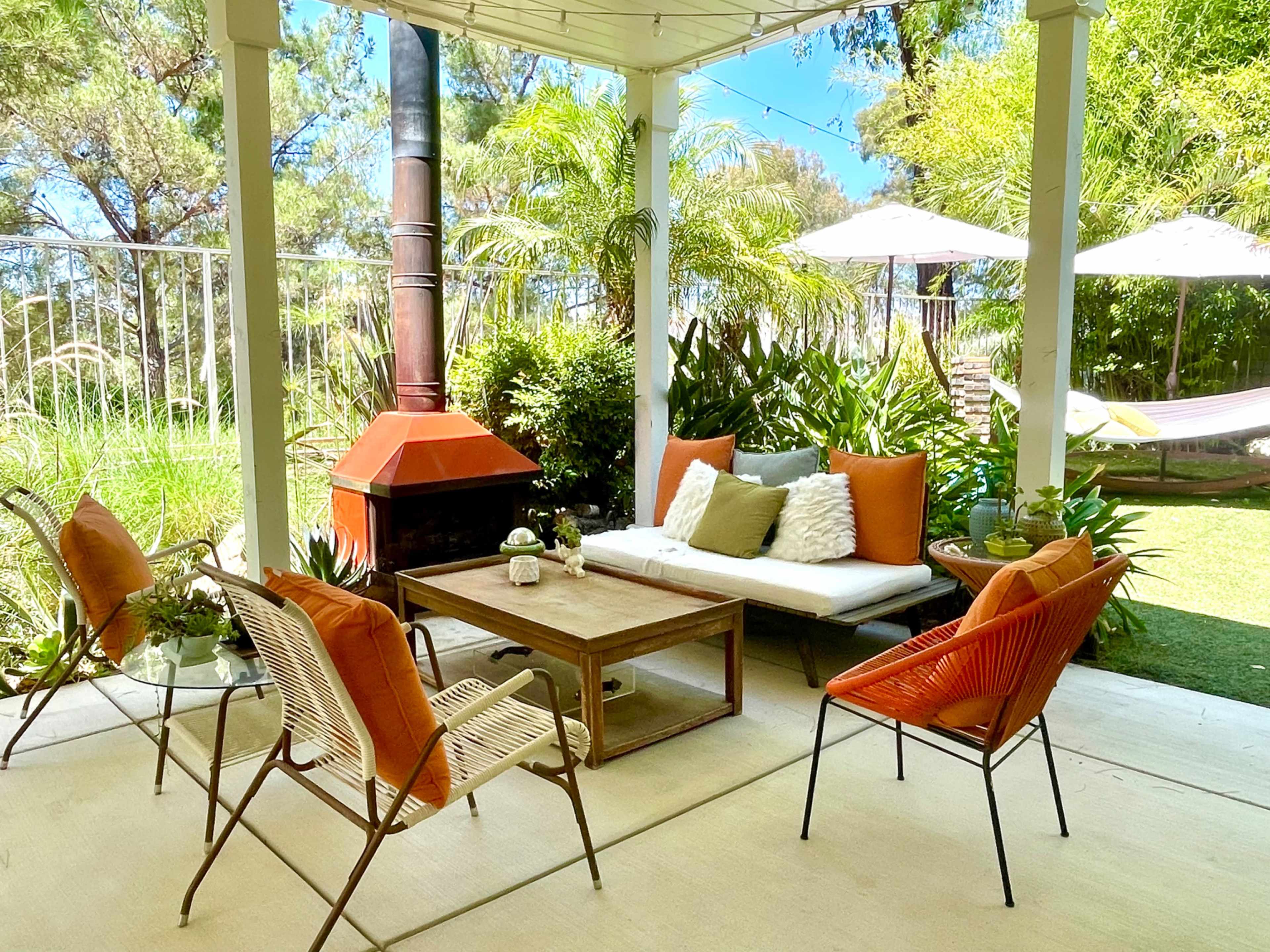 A patio area features a seating arrangement with a coffee table surrounded by chairs, a potted plant, and a fireplace, set in a landscaped garden with hammocks.