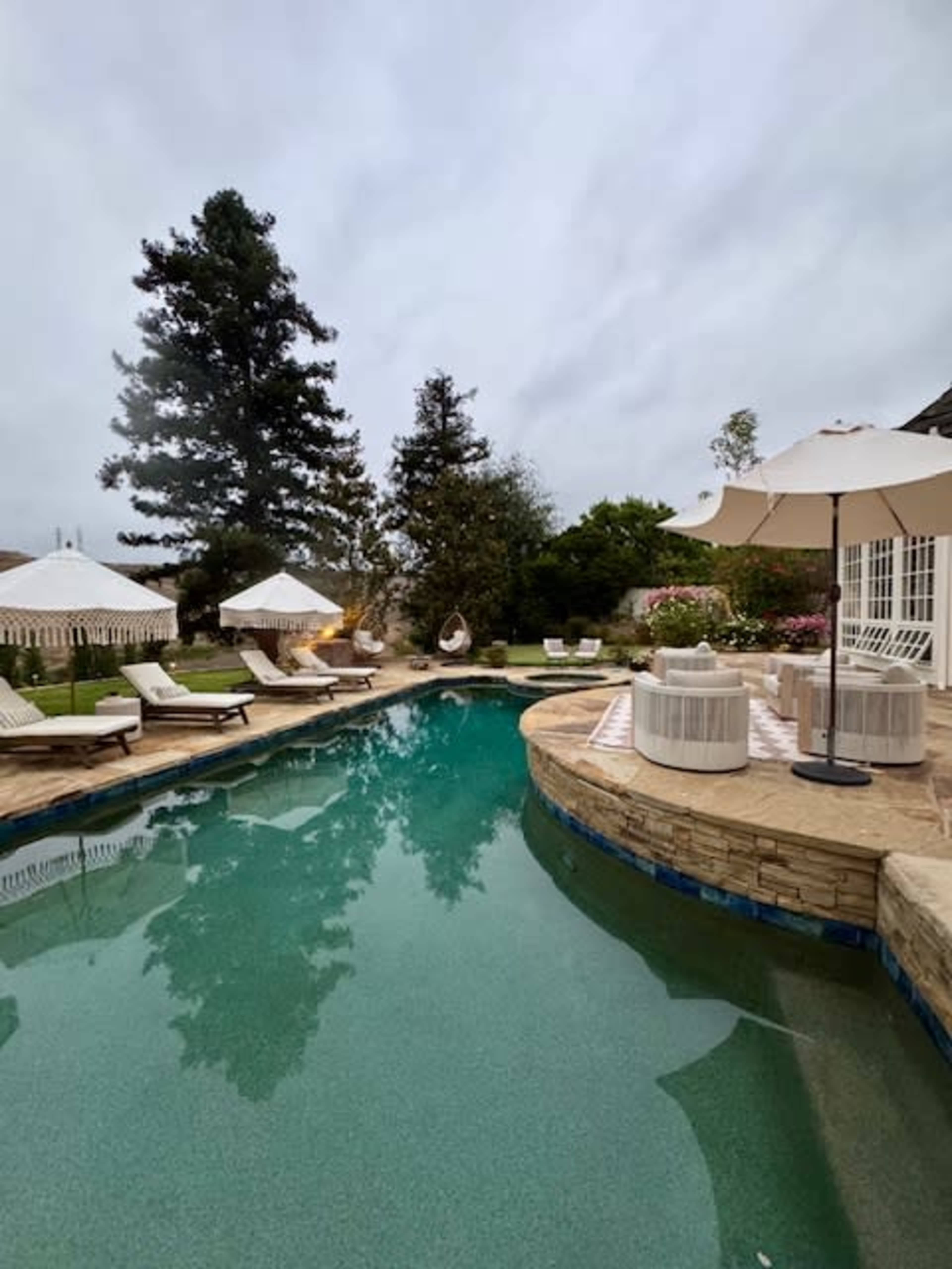 The image shows a landscaped pool area surrounded by lounging chairs, umbrellas, and greenery.