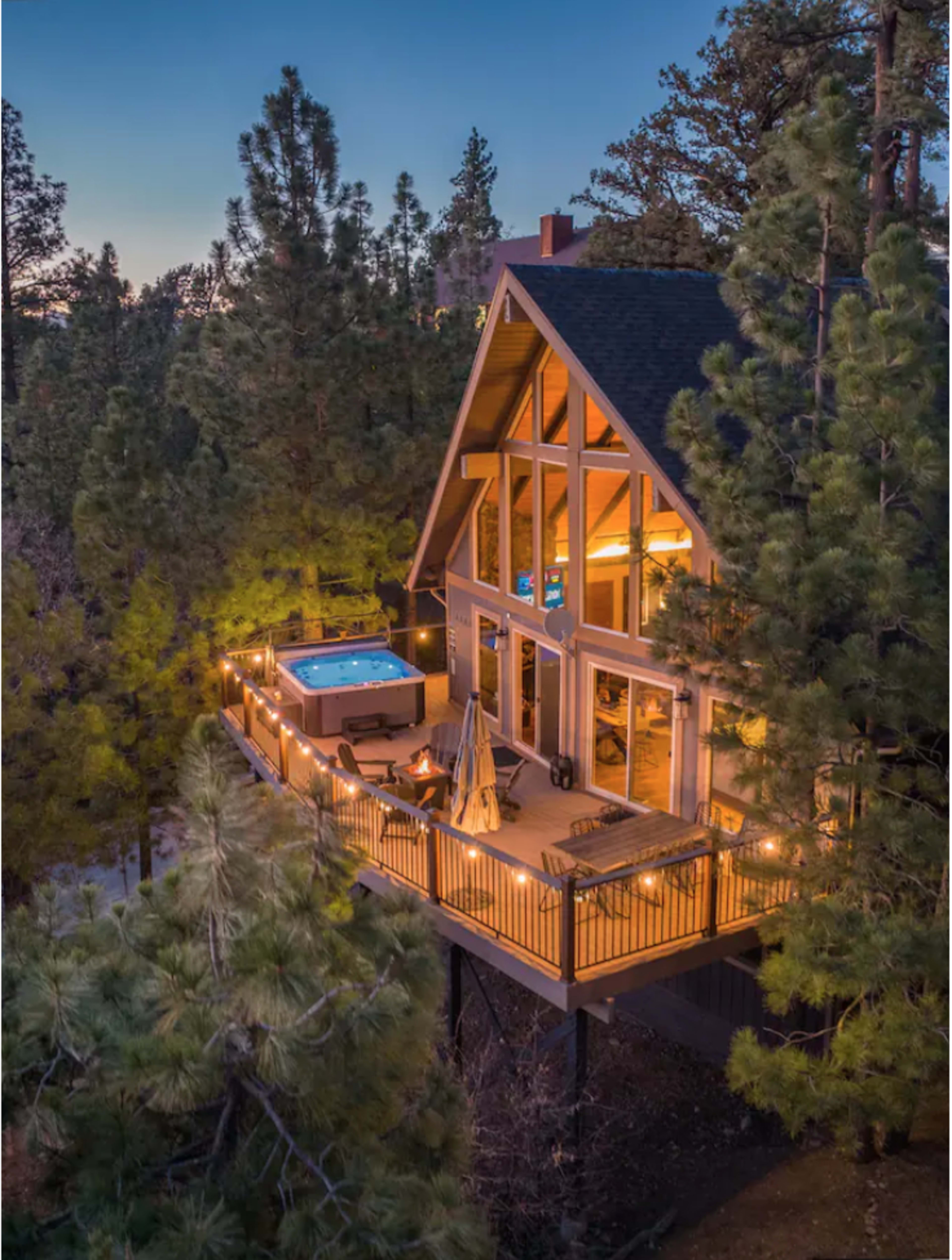 A modern cabin with large windows and a deck featuring a hot tub is surrounded by tall pine trees in the twilight.