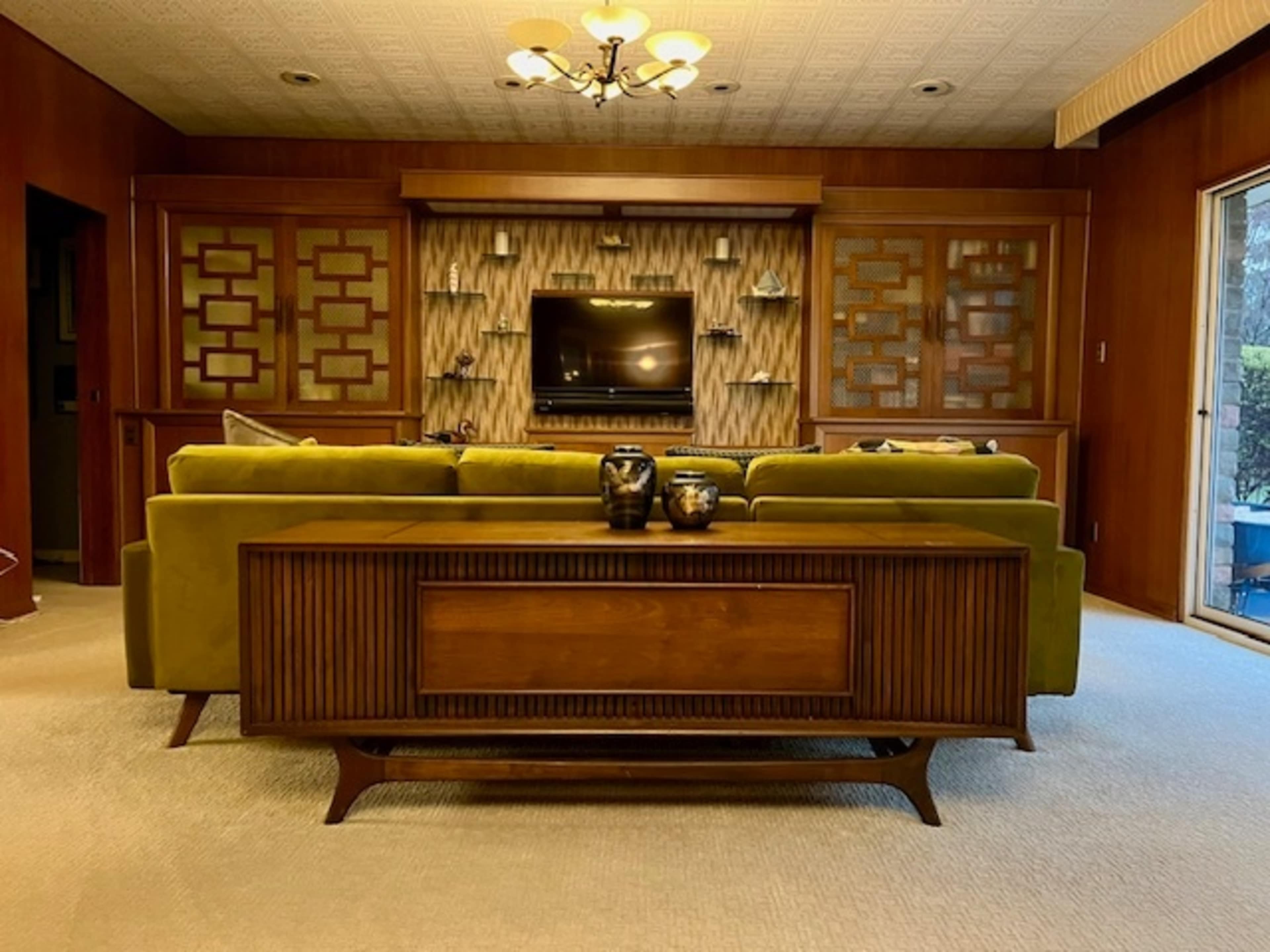 The image shows a living room with a green couch facing a wall-mounted television, surrounded by wooden cabinetry and large windows.