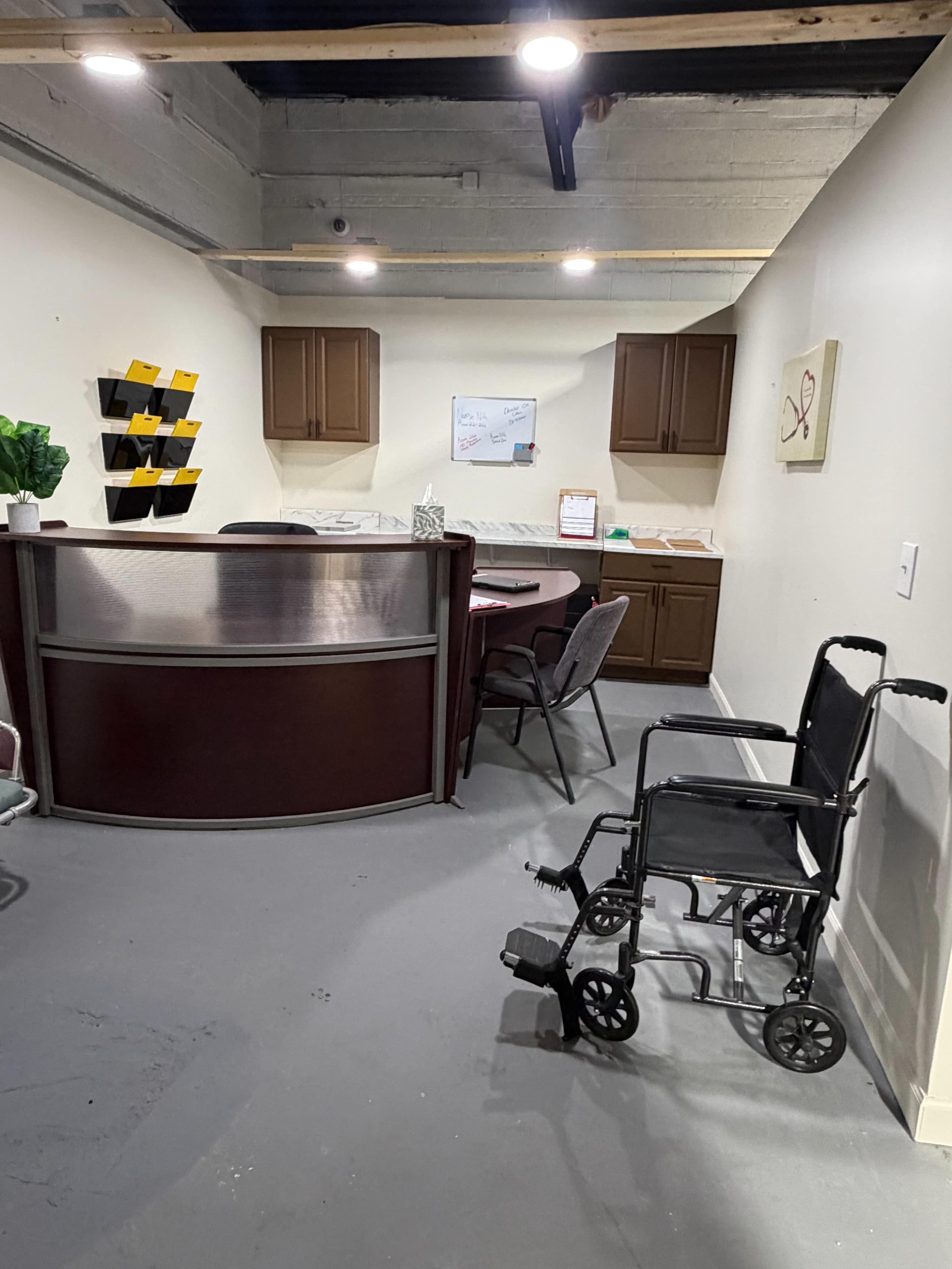 The image shows a small office or reception area with a wooden counter, chairs around a table, and a wheelchair positioned beside the wall.