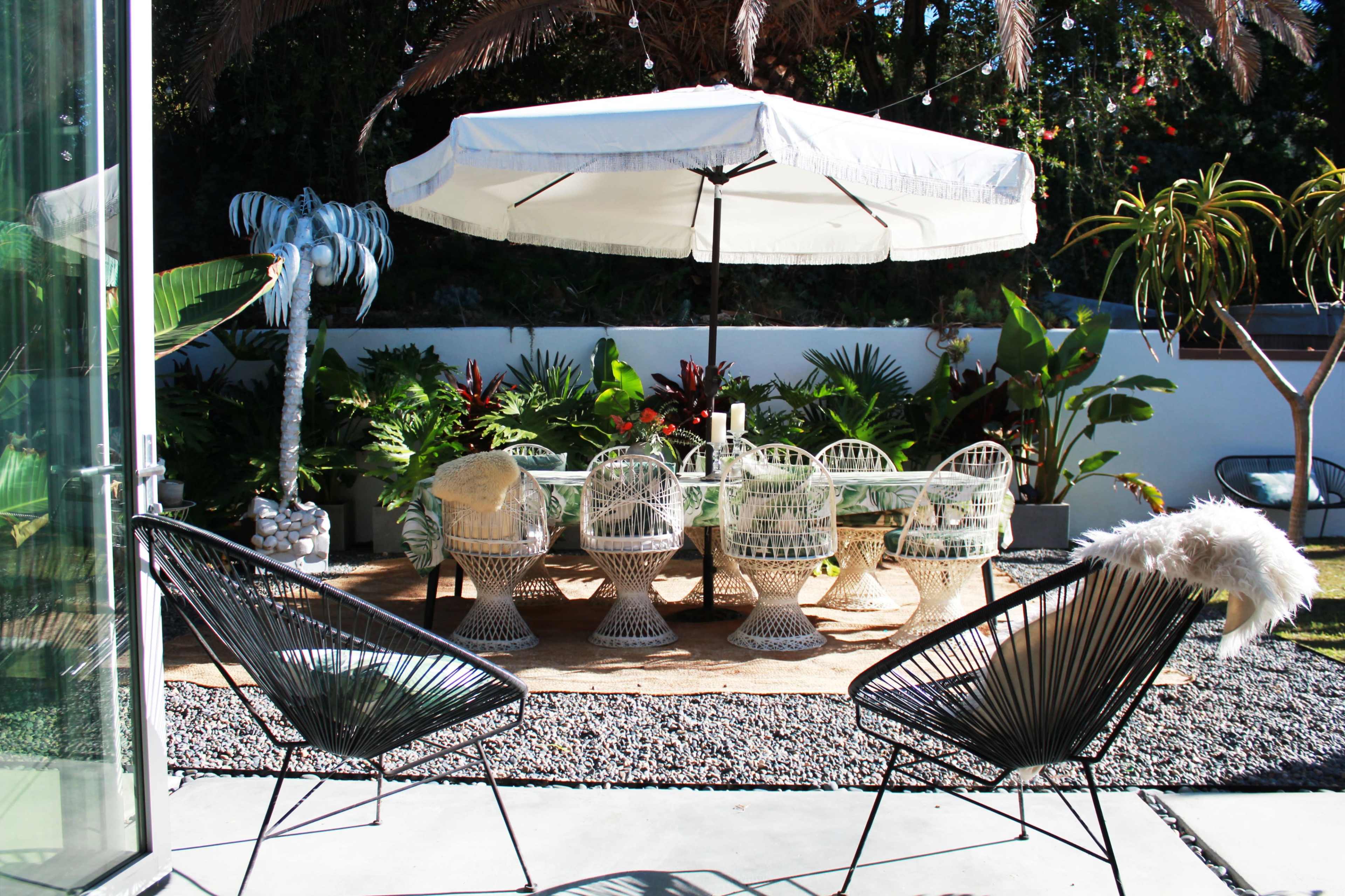 A patio area features a dining table surrounded by wicker chairs under a large umbrella, with greenery and decorative plants in the background.