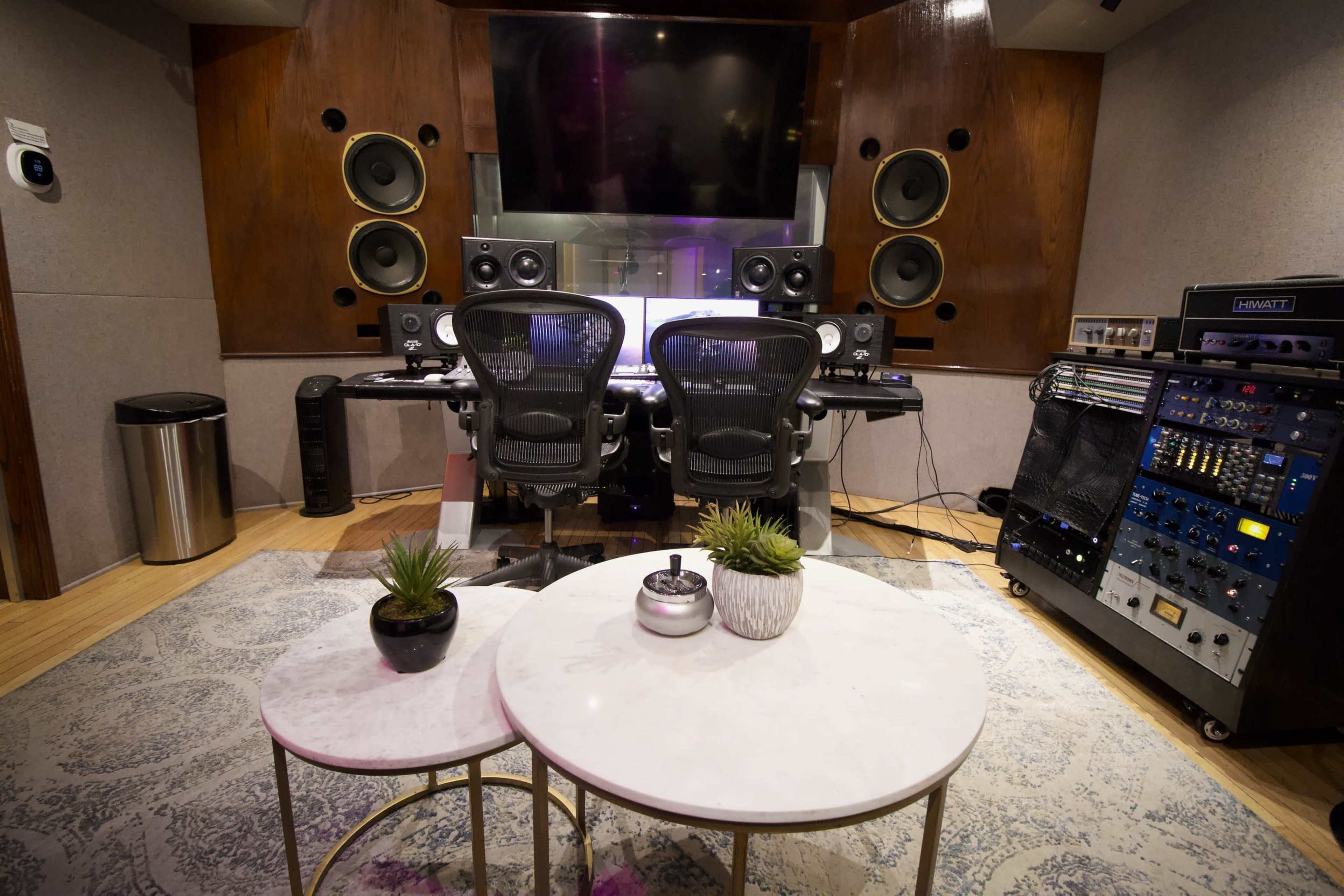A professional music studio with a mixing console, two ergonomic chairs, large speakers, and audio equipment stacked beside a small table and potted plants.