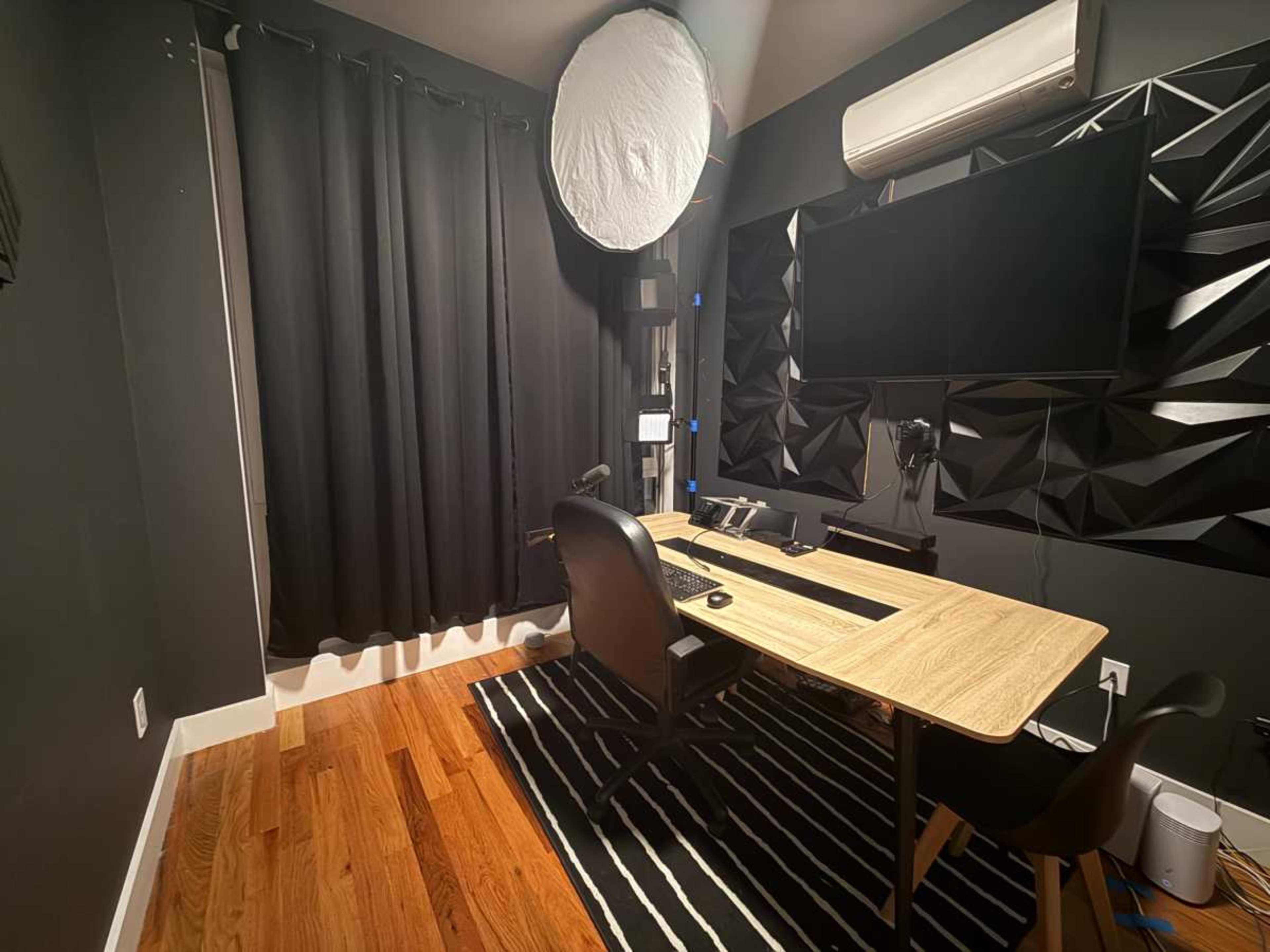 The image shows a modern home office with a wooden desk, black chair, and a wall-mounted monitor, featuring dark curtains and textured wall panels.