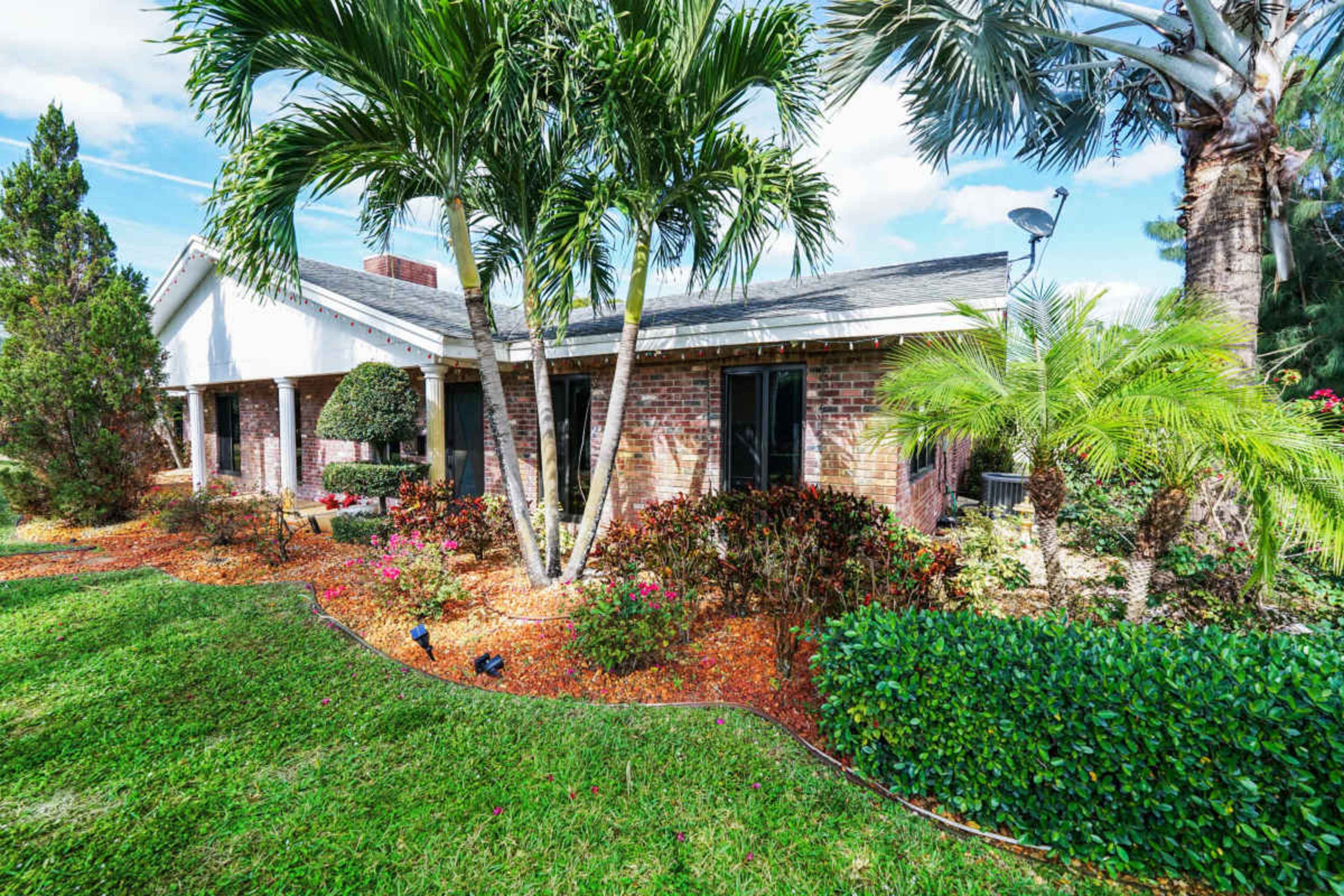 The image shows a single-story house surrounded by manicured landscaping, including palm trees, shrubs, and flower beds.