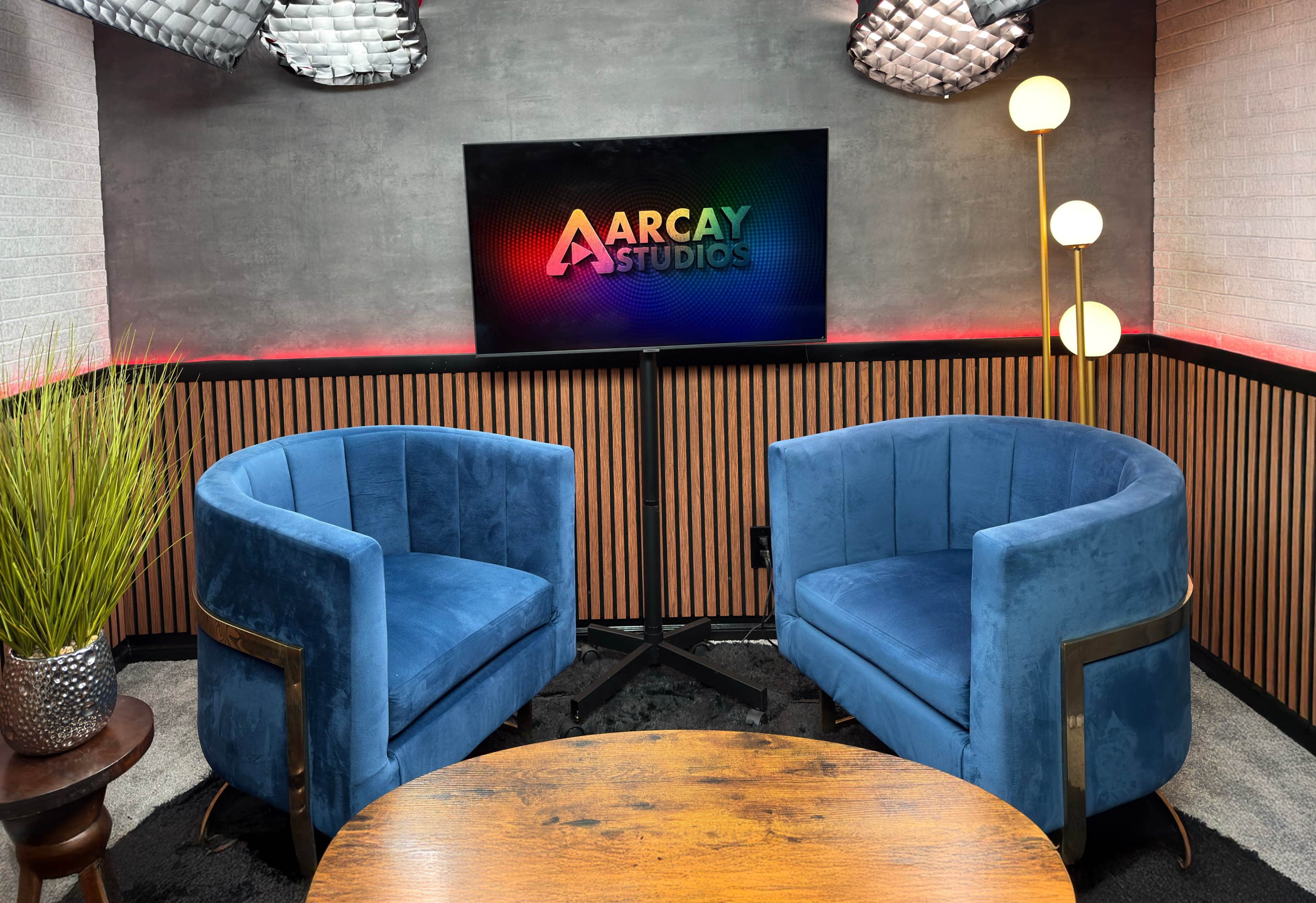 A modern studio setup features two blue velvet chairs facing a screen displaying the "Aarcay Studios" logo, with decorative elements including a plant and stylish lighting.